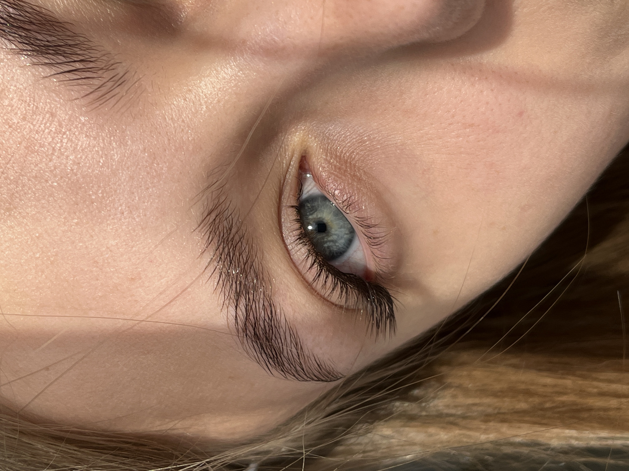 A close-up shot of a person's face angled to the side, focusing on their eye and surrounding features. The person has fair skin, and their eye is blue-gray with dark eyelashes. The eyebrows are thick and dark. There are strands of light brown or blonde hair visible near the bottom of the frame. The skin appears to have minimal makeup. The background is out of focus, but seems to be a dark or neutral color. There is no clear indication of location; the reference to Фоки, Russia, cannot be confirmed.
