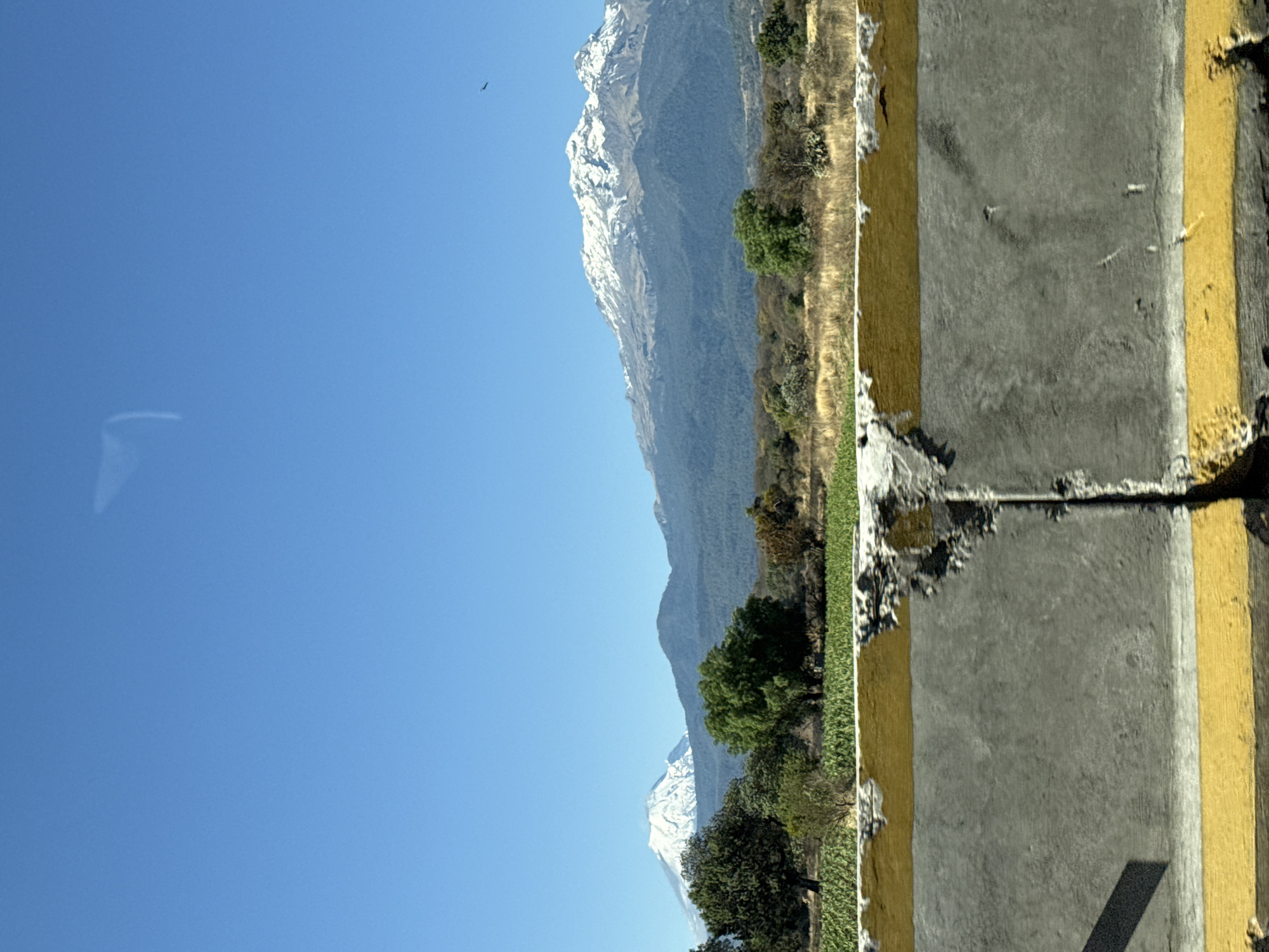 The image displays a daytime outdoor landscape located near San Matías Tlalancaleca, Mexico, under a clear, cloudless blue sky. The immediate foreground features a weathered concrete highway barrier, horizontally bisected by two prominent yellow stripes along its top and bottom edges. The concrete exhibits significant chipping, erosion, and discoloration, exposing rebar and aggregate in several areas. A distinct diagonal shadow is cast on the lower-left portion of the barrier.
Beyond the barrier, the mid-ground comprises agricultural fields with vibrant green crops, transitioning into an expanse of dry, golden-brown vegetation, interspersed with varying sizes of green trees and scattered cactus plants. The distant background is dominated by two snow-capped volcanoes. To the left, a conical peak is prominently visible with extensive white snow coverage. To the right, a larger, broader mountain range also displays significant snow on its upper slopes, with its lower elevations covered in dark green forest. Multiple layers of forested hills are visible between the foreground and the major peaks. A small, dark, unidentified flying object is present in the upper right quadrant of the sky. Reflections, consistent with an interior window, are visible on the upper portion of the image frame.