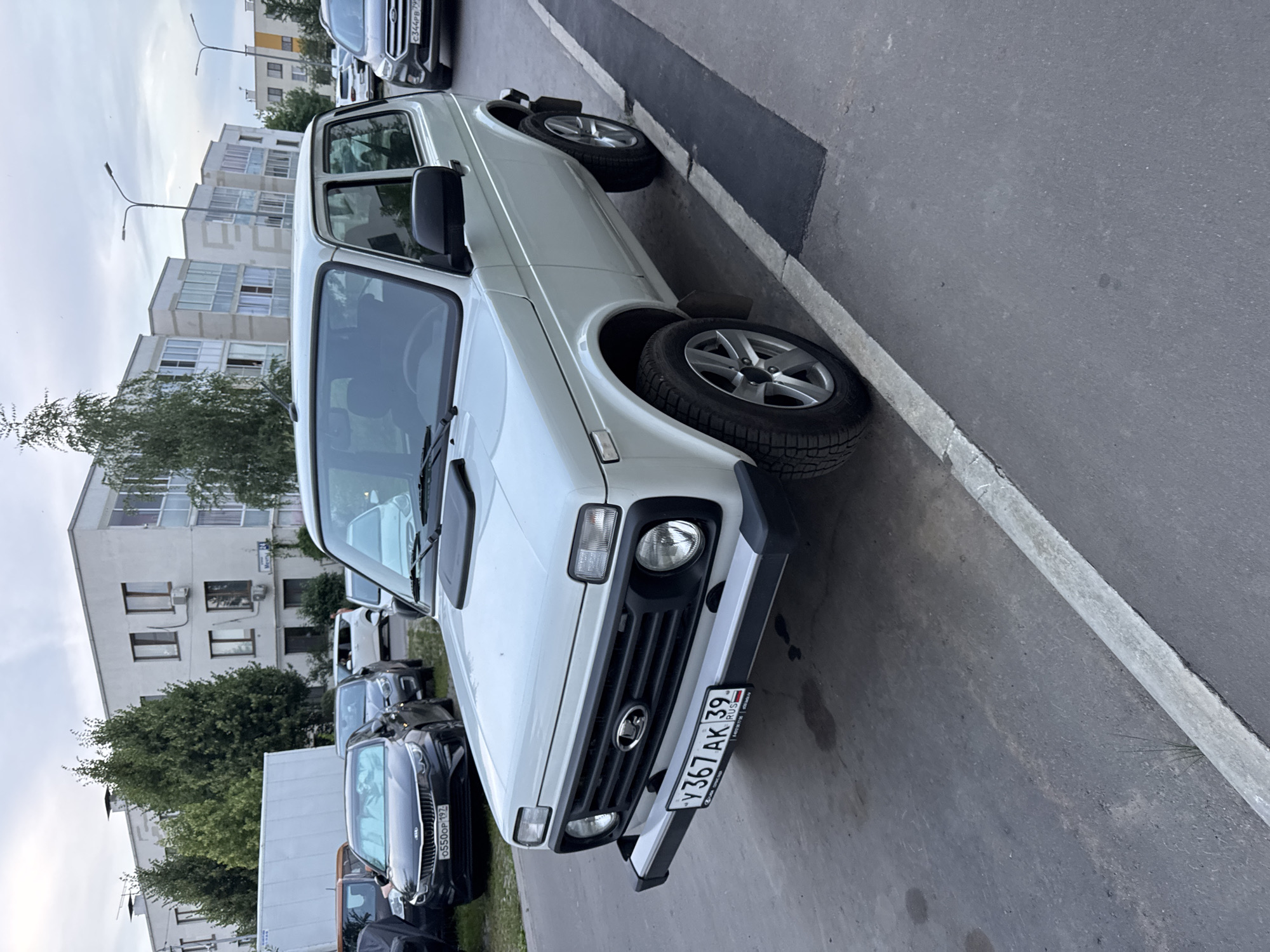 The image depicts a white Lada Niva vehicle parked on an asphalt surface next to a concrete curb in an urban environment. The vehicle is positioned at an angle, with its front facing slightly towards the viewer. The car has a standard Lada emblem on the front grille and a Russian license plate reading "Y367AK 39". In the background, several multi-story apartment buildings with light-colored facades and balconies are visible. Trees and streetlights are also present. Other vehicles, including a black Kia sedan with a Russian license plate "0550OP 197", are parked behind the Lada Niva. The sky is overcast, indicating a cloudy day. The location is identified as Domodedovo, Russia, suggesting this is a residential area.