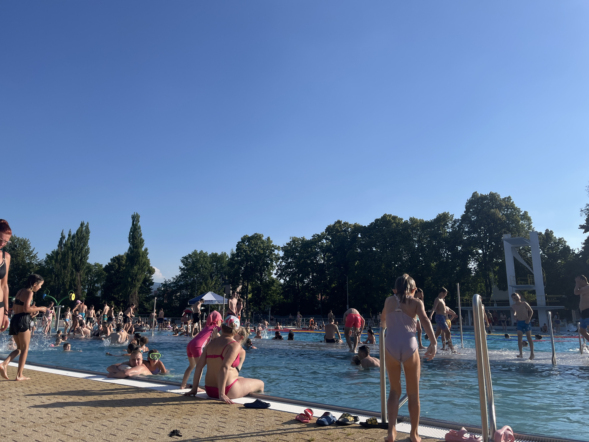 The image depicts a bustling outdoor swimming pool scene on a bright, sunny day, likely in the afternoon given the warm light and long shadows. A clear blue sky dominates the upper portion of the frame, with lush green trees forming a backdrop along the perimeter of the pool area.
The pool itself is filled with a large number of people, appearing to be a mix of adults and children, all engaged in various water-related activities. Some are swimming, others are standing or sitting at the edge of the pool, and a few seem to be playing or splashing in the water. The water is a clear blue, and the pool features a diving board structure in the background. The pool deck is made of a textured, light-colored material, and several pairs of flip-flops are scattered near the edge.
In the foreground, a young girl in a light purple swimsuit is facing away from the camera, looking towards the pool. To her left, a woman in a pink bikini is seated on the edge of the pool. The overall atmosphere appears to be one of recreation and enjoyment, with people of various ages enjoying the warm weather and the cool water. The presence of numerous individuals suggests a popular public pool or a resort. No discernible text is visible in the image.