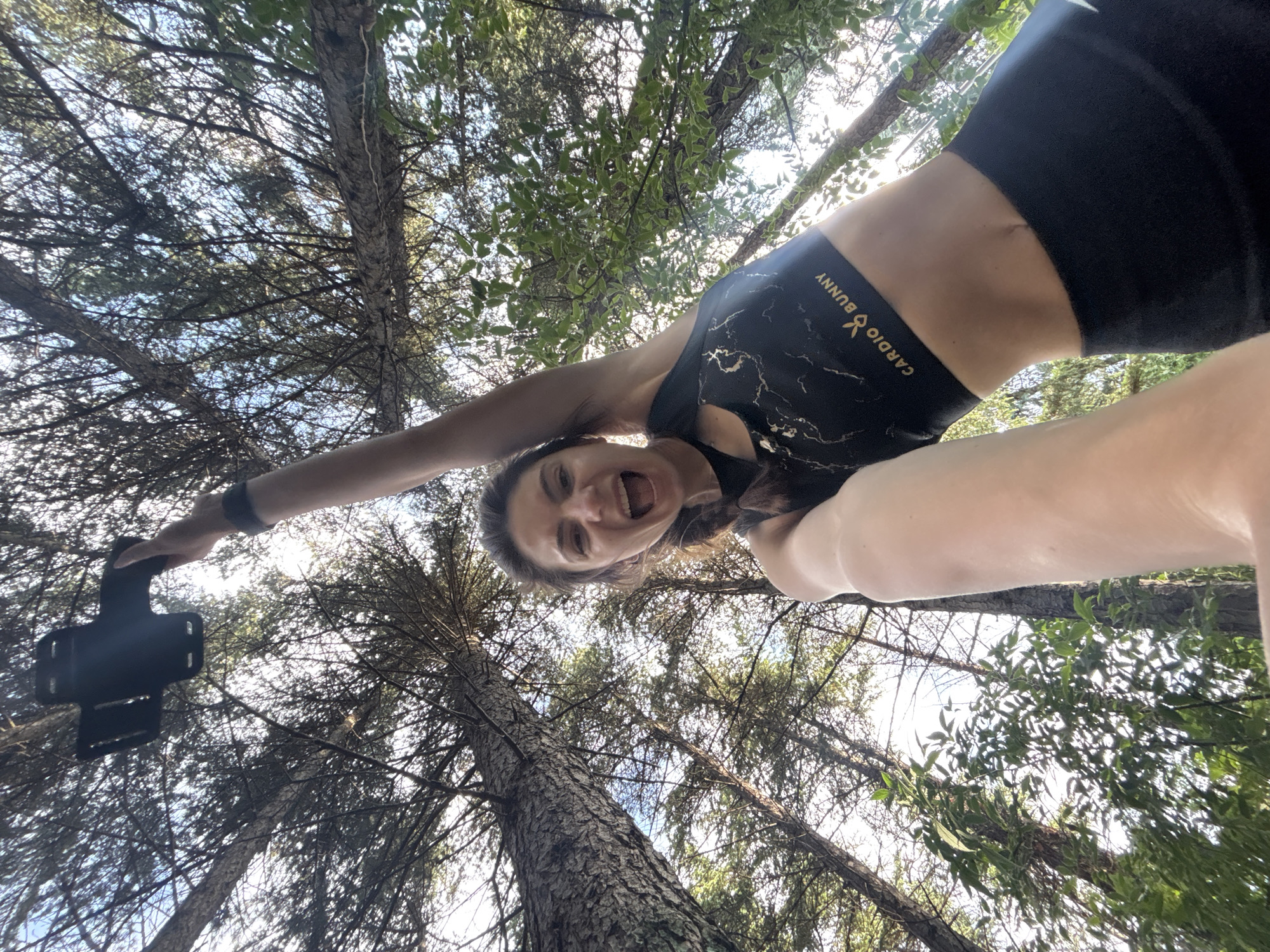 A woman is taking a low-angle selfie in a forest. She is wearing a black sports bra with a gold logo that says "CARDIO BUNNY" and black leggings. Her right arm is extended upwards, holding a black object, possibly a phone mount. She has brown hair braided to her side and is smiling with her mouth open. The background is filled with trees and a bright sky. The scene appears to be in a natural setting, possibly a park or hiking trail within the city of Varna, Bulgaria.