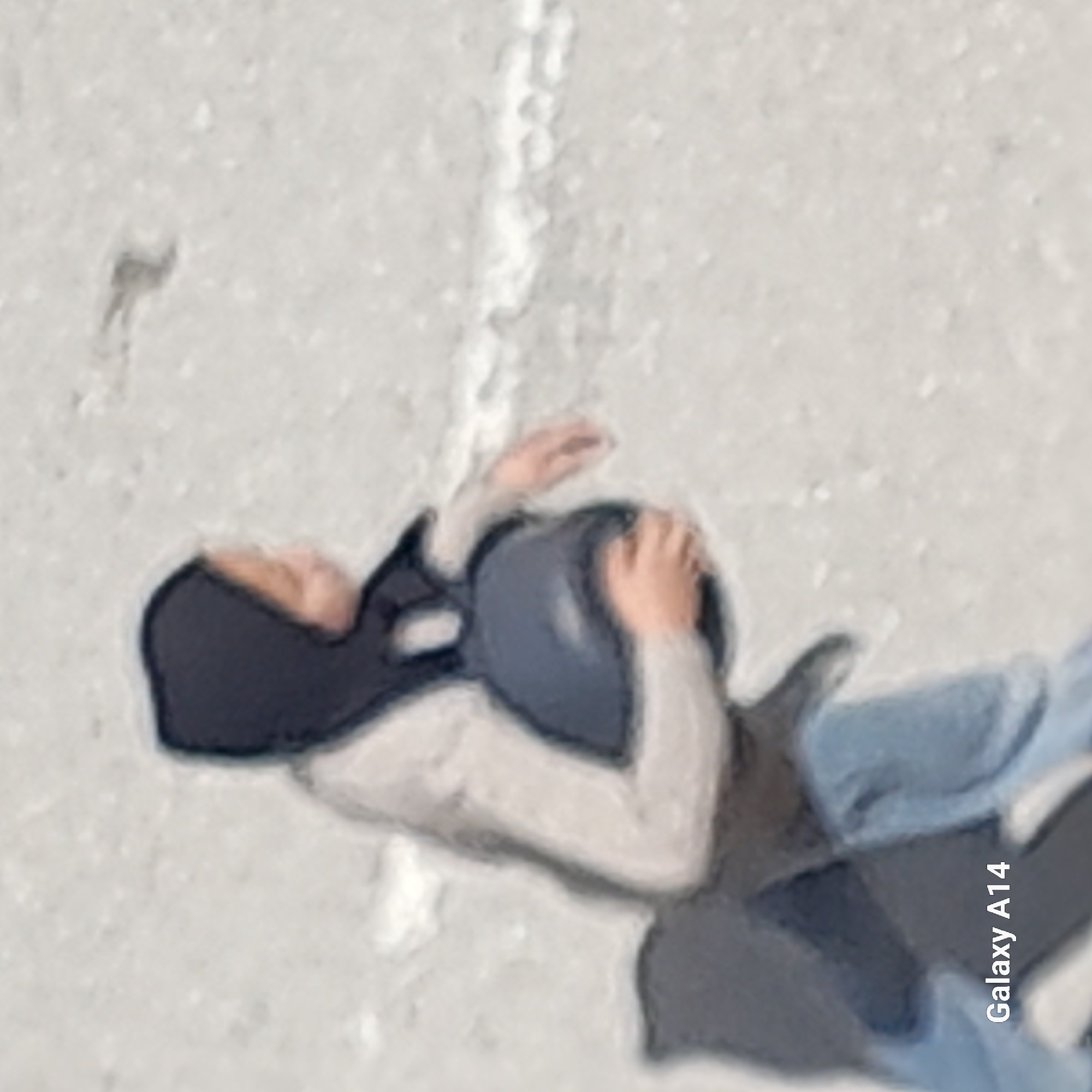 A person wearing a dark head covering and blue jeans appears to be lying on a light-colored, possibly snow-covered, surface. They have their hands raised near their head, which is positioned close to a dark object. A thin line is visible on the surface in the background. The image appears to be a cell phone photo, based on the "Galaxy A14" text in the corner. The specific location within the city of Unknown, Unknown is not identifiable from the image.
