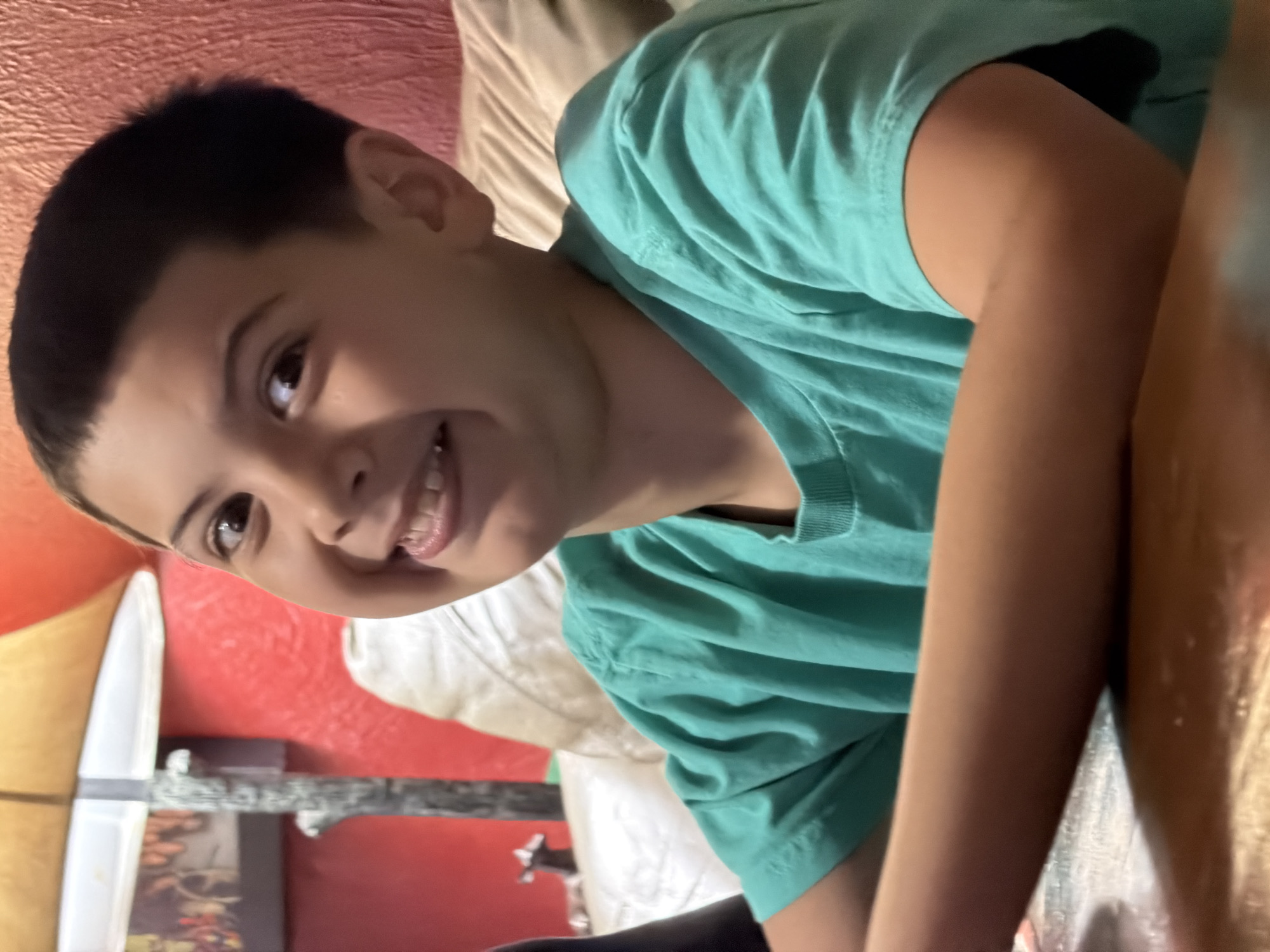 A young boy, with dark hair and a wide smile, is photographed from the chest up. He is wearing a teal t-shirt and is seated at a wooden table. Behind him, a textured red wall and a portion of a cream-colored couch are visible. A lamp with a tan shade is also present. The scene appears to be an indoor residential setting. The image does not contain enough information to determine a specific location within Virginia Beach, United States of America.
