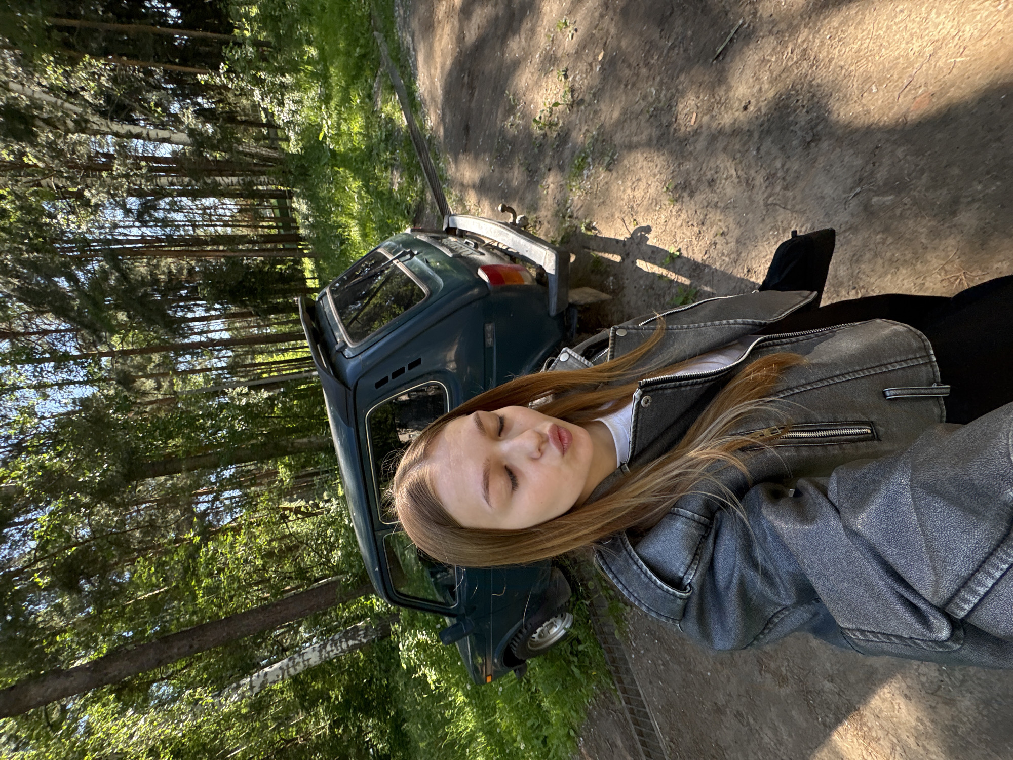 In a sun-dappled forest clearing, a young woman with long, light brown hair takes a selfie. She is wearing a gray faux-leather jacket over a white t-shirt and dark pants, and she is puckering her lips as if for a kiss. To her left, a dark green, older model SUV, possibly a Lada Niva, is parked on a dirt road. The car is surrounded by lush green grass and the dense trees of the forest, typical of the natural landscapes found near Chaikovsky, Russia. The ground is covered in dirt and scattered leaves, with sunlight casting long shadows. A metal grate is visible in the foreground.