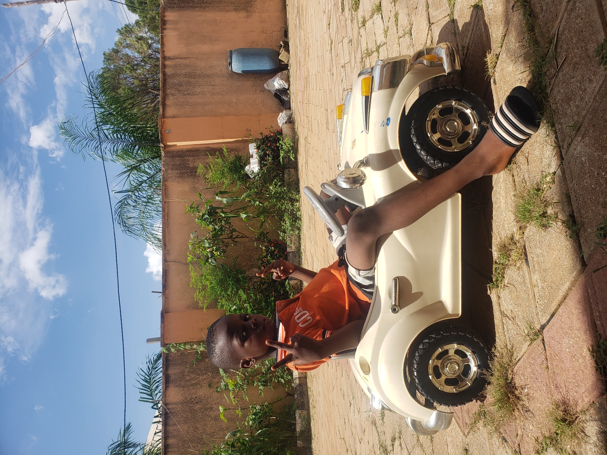 A young Black boy, appearing to be around 6-8 years old, sits in a cream-colored toy car. He is wearing an orange t-shirt with white writing and gray and white striped shorts. He is looking at the camera with a neutral expression and holding up two fingers on each hand, forming peace signs. The toy car has a steering wheel, chrome accents, and yellow headlights. The boy is wearing black and white striped slides on his feet. He is positioned on a paved patio with square concrete tiles interspersed with patches of grass. Behind the boy and the car is a peach-colored stucco wall. Green foliage is visible growing against the wall and in the background. A tall blue plastic barrel and some white and grey bags of refuse are also against the wall to the right. The sky above is blue with scattered white clouds. The scene is outdoors, suggesting a residential area in Sabon Tasha, Nigeria.