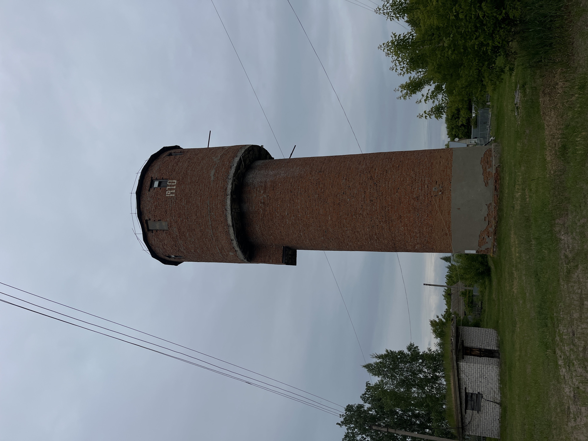 The image depicts a tall, cylindrical brick tower in Novocheremshansk, Russia, under an overcast sky. The tower, constructed of red brick, features the year "1910" prominently inscribed in white numerals on its upper section. This section, wider than its base, includes small rectangular window openings and is topped with a narrow railing. A concrete ledge separates the upper and lower cylindrical sections. The tower's base shows deterioration, with concrete plaster peeling to expose bricks.
To the left of the tower, a small, single-story building of white cinder blocks or bricks is visible. Its flat roof is covered with green vegetation. The building has a dark, possibly boarded-up window opening and a dark wooden door on its visible side. The ground is covered with green grass, with some patches of bare earth.
Overhead, multiple dark power lines extend diagonally across the gray sky from the upper left. Various trees are present: taller deciduous trees with lighter green foliage on the left, and smaller, bushier trees with darker green leaves on the right. In the distance, behind the smaller building and trees, hints of other structures and a wooden fence are discernible. No people or animals are visible in the scene.