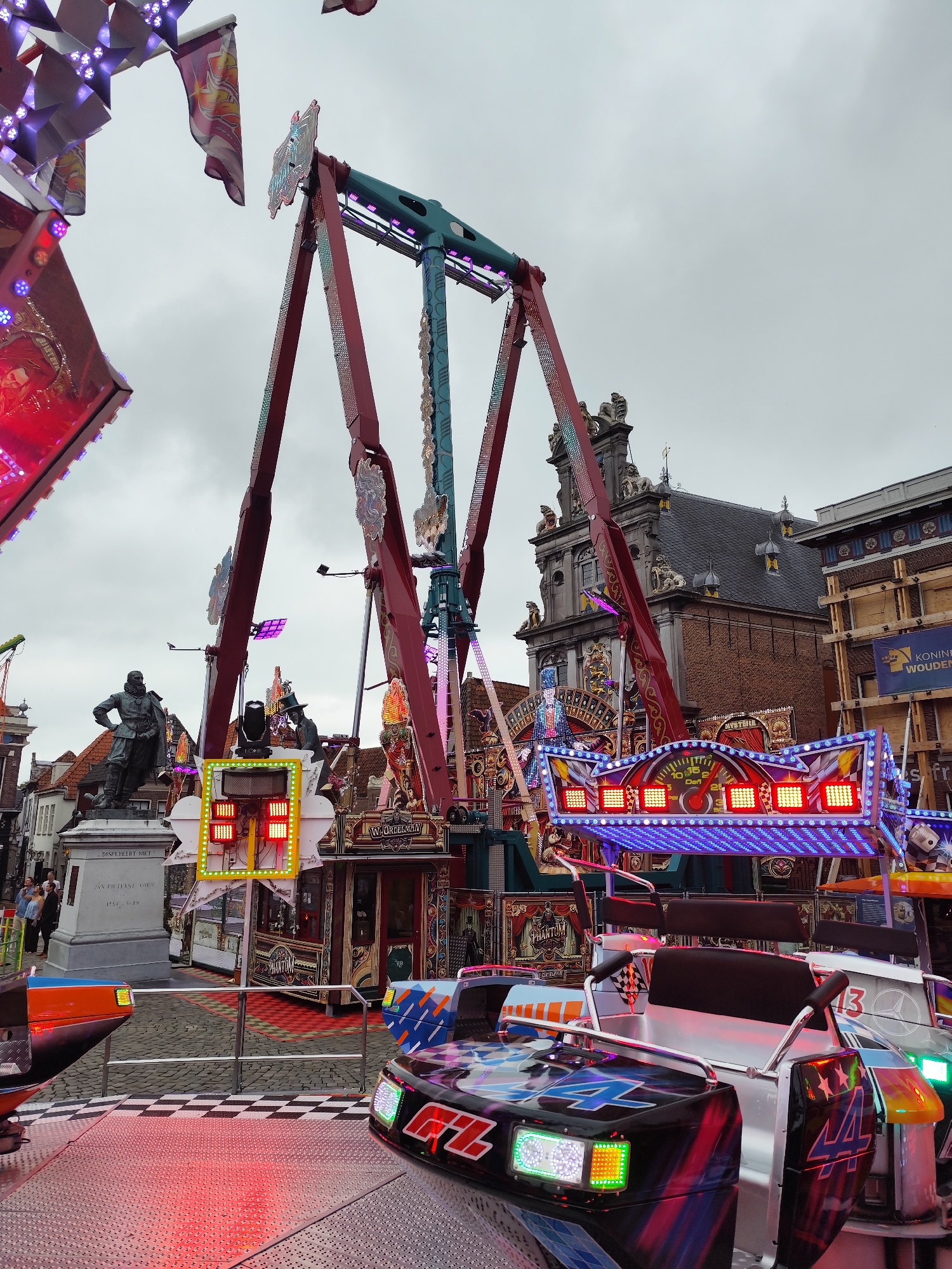 In the city of Hoorn, Netherlands, an amusement park scene unfolds under a cloudy sky. The central focus is a large pendulum ride with red and teal structural components. The ride is adorned with colorful lights, primarily red and blue, that illuminate the surrounding area. In the foreground, parts of a smaller ride or attraction are visible, featuring metallic surfaces and vibrant decals. One car-like seat in the foreground has "44" emblazoned on it. Behind the rides, a statue of a man in historical attire stands on a pedestal. The pedestal has inscriptions that appear to be in Dutch. A building with a traditional Dutch architectural style, featuring a dark tiled roof and ornate facade, is visible in the background. Other structures with signage indicating commercial activity are also present. A few individuals can be seen walking in the mid-ground, appearing as silhouettes against the rides. The ground is paved with cobblestones and a checkered pattern.