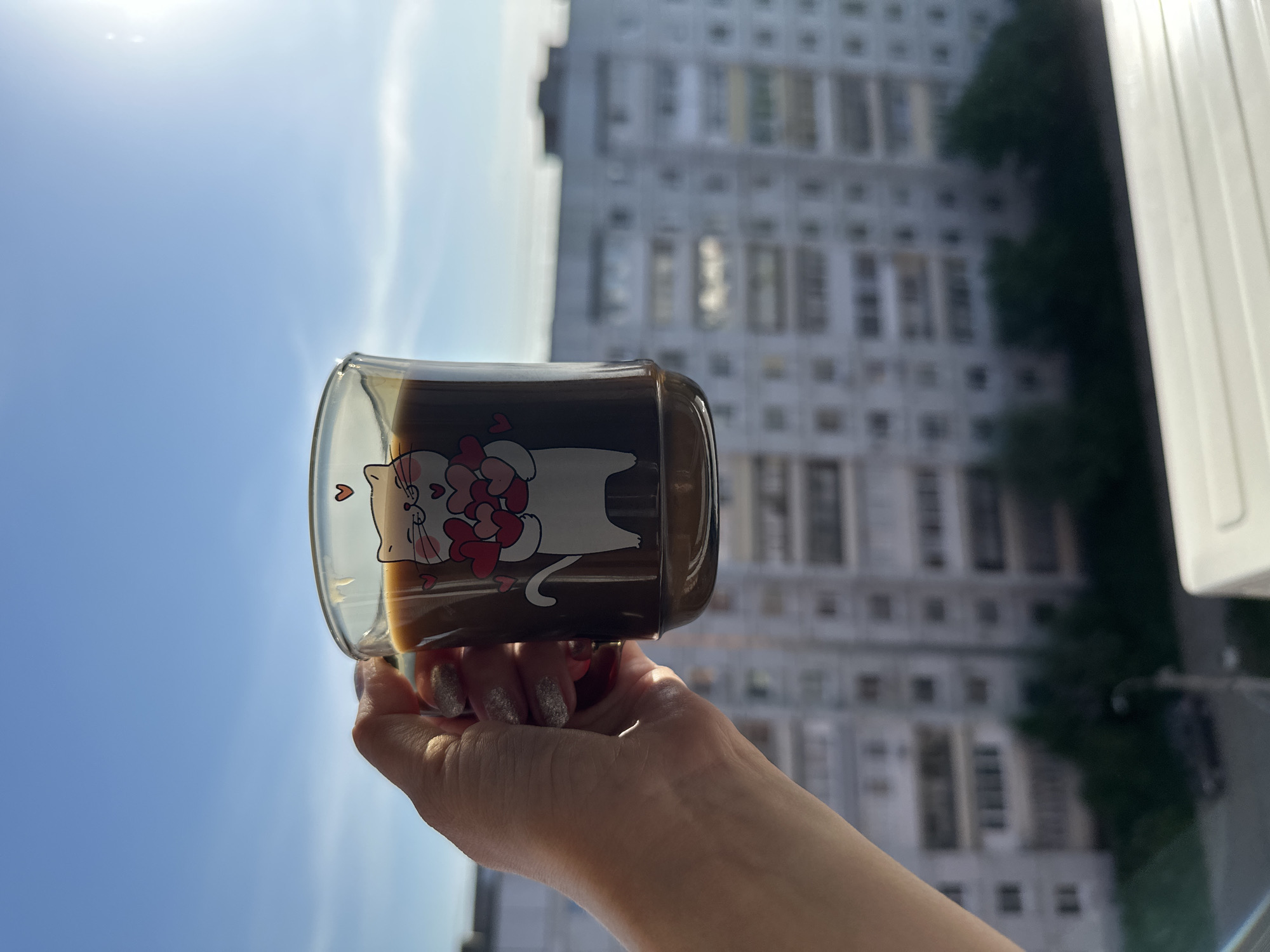 A hand holds a glass mug with a cartoon cat design, containing a dark liquid. The backdrop features a light blue sky and a multi-story building with numerous windows. The location is Chaikovsky, Russia.