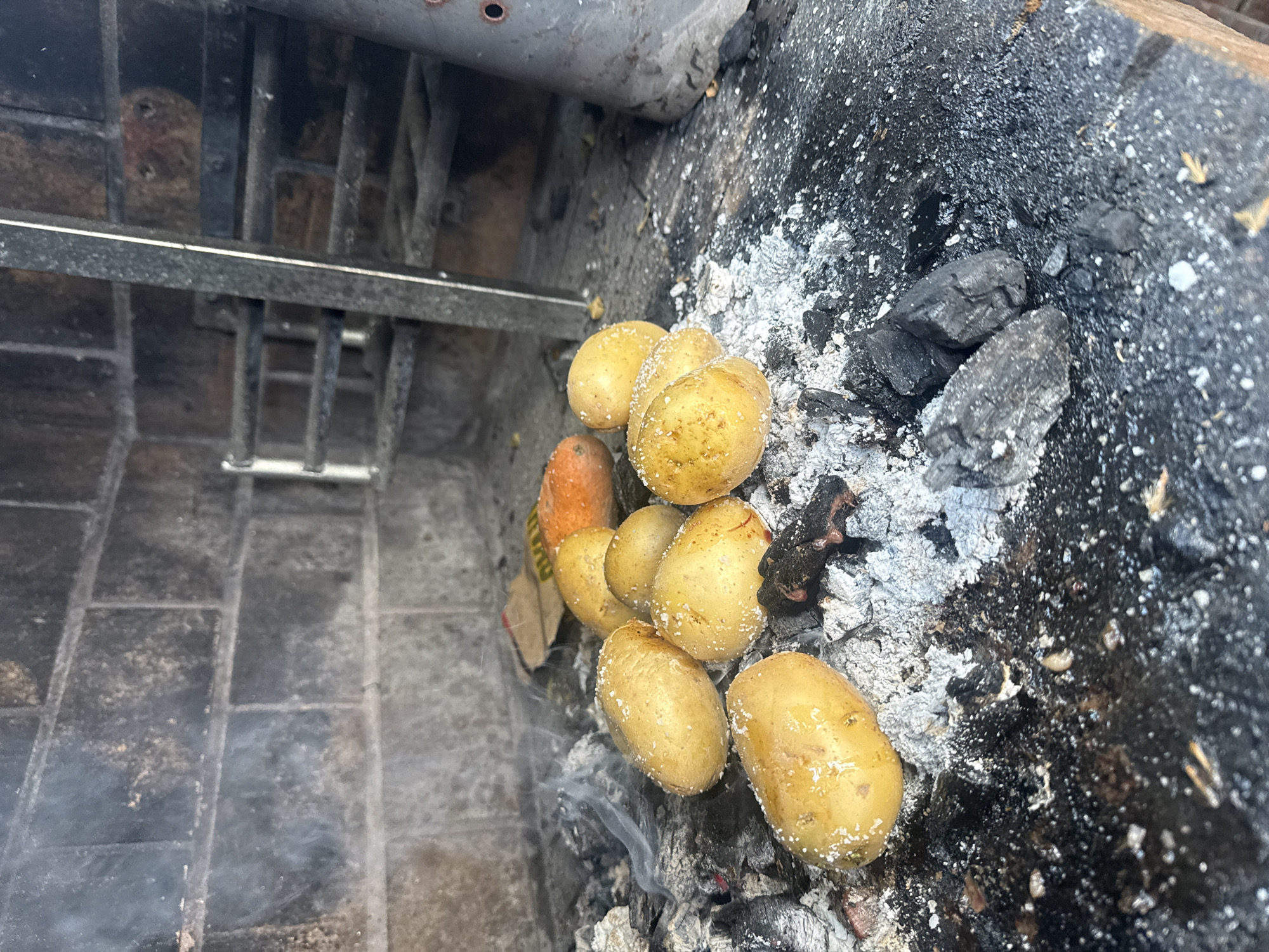 The image shows a close-up of potatoes roasting in a fireplace or fire pit. The potatoes, coated with what appears to be salt, are nestled among hot coals and ash. There is also an orange object among the potatoes, possibly a carrot. A metal grate or support structure is visible in the background, situated near brickwork. The overall environment suggests an outdoor or semi-outdoor setting, possibly a backyard or patio. The location in the city of Barcelona, Spain cannot be confirmed based on the image alone.