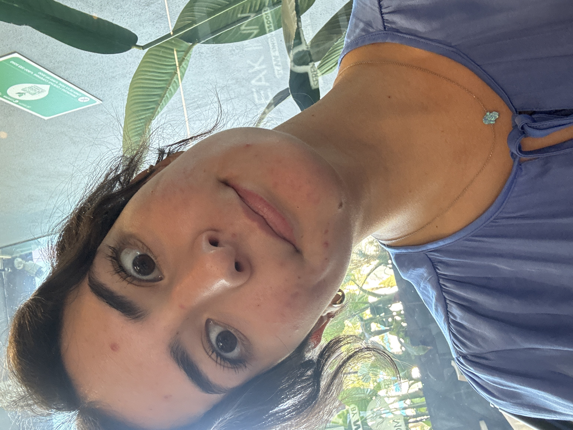 The image is a close-up selfie of a young woman with dark hair and brown eyes. She has some minor skin blemishes on her face. She is wearing a light purple top and a delicate necklace with a blue pendant.
In the blurred background, hints of plants and signage are visible. The signage reads "FRANKEN", suggesting the location is possibly a restaurant or business with a similar name. Further text on the sign includes "Gran Aeropuerto". Based on this information, it is likely that the photo was taken at or near the Puebla International Airport (officially known as Aeropuerto Internacional de Puebla "Hermanos Serdán"), located in Puebla, Mexico.