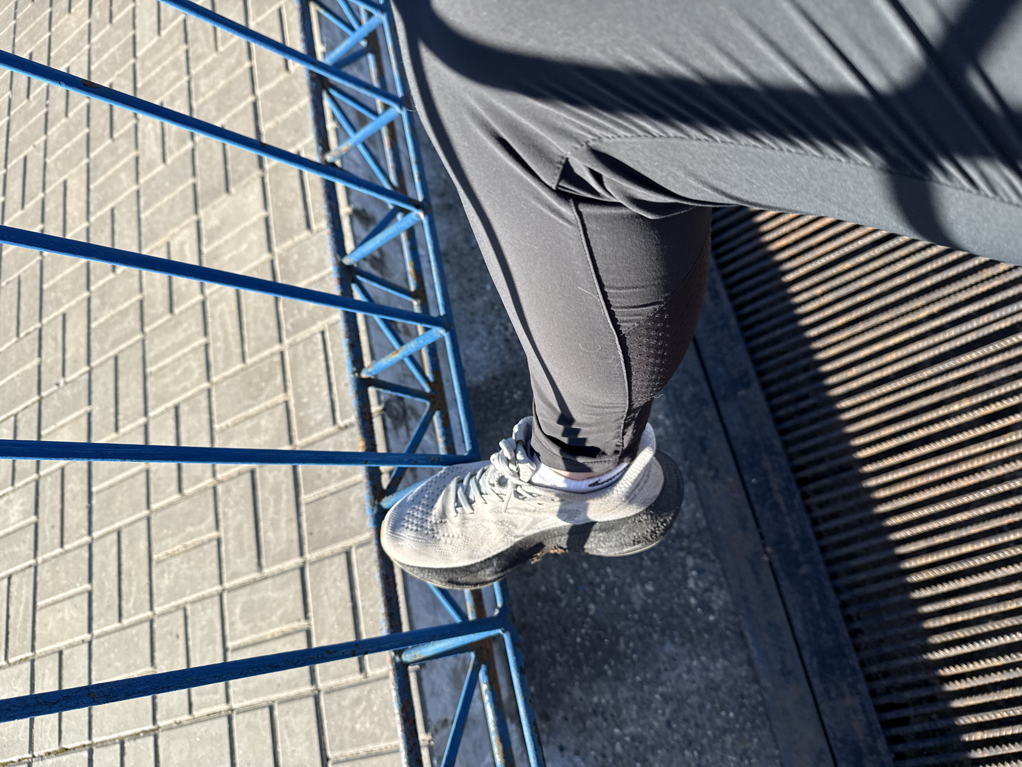 The image depicts a person standing on a metal grating. The person is wearing black athletic pants and a gray sneaker with a white sock. A blue metal railing is to the left, with a stone-block walkway beyond. A rusted metal grating is to the right. The location is the city of Chaikovsky, Russia.