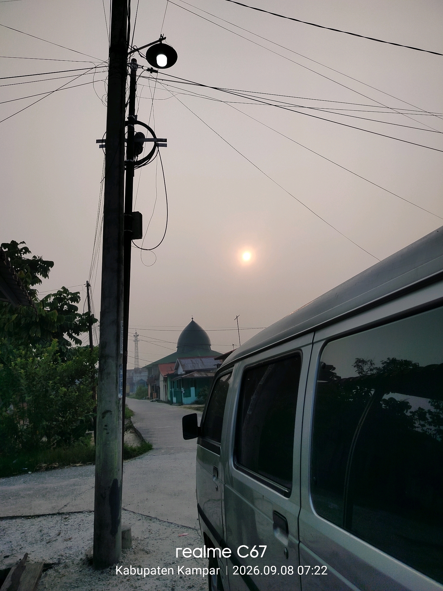 This image captures an outdoor scene in Kubang Jaya, Indonesia. In the foreground, a light-colored van is parked on the right side of a paved road. The van's windows reflect the hazy sky and some surrounding foliage. To the left of the van, a utility pole stands tall, adorned with numerous electrical wires and a lit streetlamp. Beyond the pole, a road stretches into the distance, flanked by houses and lush green trees. Further down the road, a mosque with a green dome is visible. The sun, partially obscured by haze, hangs in the sky, casting a soft glow over the scene. The image is timestamped with "2026.09.08 07:22" and tagged with "realme C67" and "Kabupaten Kampar".
