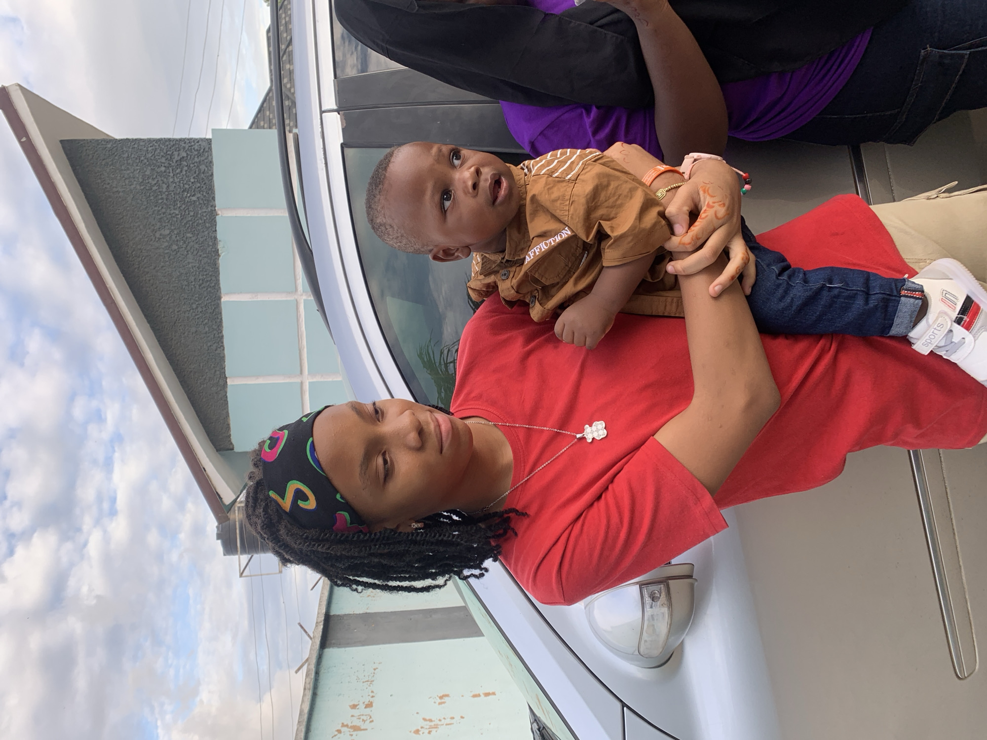The image depicts an outdoor scene in Jos, Nigeria, featuring three individuals positioned in front of a silver vehicle and two distinct buildings under a partly cloudy sky.
On the left, a young female, estimated to be a teenager or young adult, stands holding a toddler. She has dark skin and long, dark hair styled in braids, secured by a black headband with multi-colored, abstract patterns. She wears a plain red short-sleeved t-shirt, a silver necklace with a teddy bear pendant, and small stud earrings. Her gaze is directed downwards and to her right. Her visible left hand has short, unpainted nails. She is wearing light-colored (possibly khaki) shorts or trousers, and white sneakers with red, black, and white accents, displaying the word "sports."
She holds a male toddler, approximately 1-3 years old, with dark skin and short hair. The child wears a light brown long-sleeved collared shirt with "AFFLICTION" printed on the left sleeve and white stripes on the right sleeve. He is dressed in dark blue jeans. His left hand exhibits orange-red henna-like markings, and he wears a thin orange bracelet on his left wrist and a pink bracelet on his right wrist. The child's eyes are directed towards his right, with a neutral expression.
To the right of the child, a third individual is partially visible. This person is an adult, wearing a purple top and dark jeans or trousers. A black hijab or head covering is draped over their head and shoulder, obscuring most of their face and upper body. This individual's right arm is also supporting the toddler.
In the background, a silver sedan or SUV is parked. Its right side mirror is visible, along with reflections of the sky and vegetation on its windows. Behind the vehicle and individuals, two distinct buildings are present. To the left, a light blue wall or section of a building is topped with a barbed wire fence. To the right, a multi-story building features a dark grey, textured upper section with a pointed roofline trimmed in brown. Below this is a light blue tiled or painted section, followed by another dark grey textured segment. A black water tank on a stand is visible above the roofline of the left background building section. The sky overhead is partially covered by white clouds, with areas of blue sky visible.