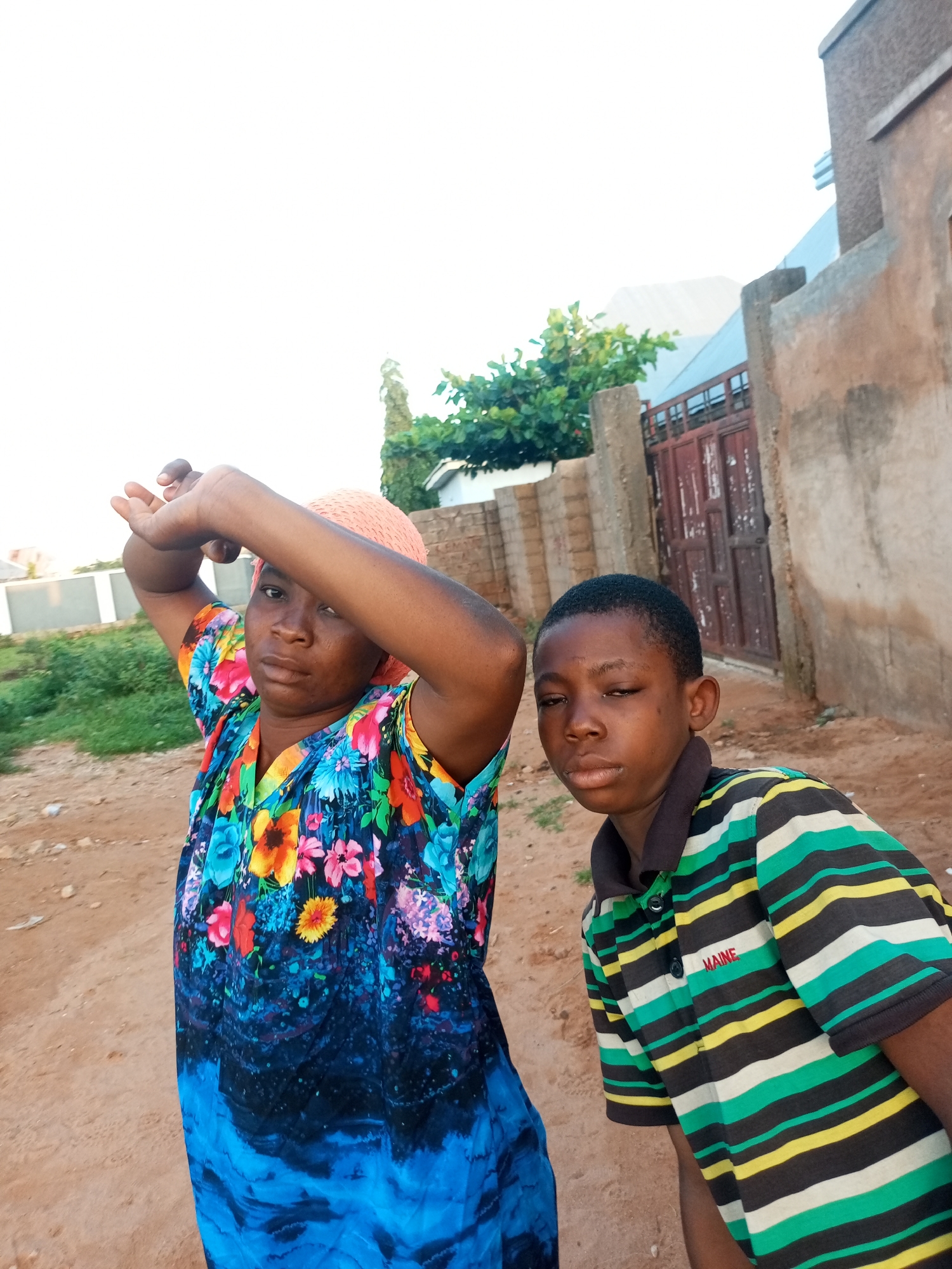In Tempure, Nigeria, two individuals, an adult female and a young male, are captured in an outdoor, low-angle portrait. The female, positioned on the left, wears a vibrantly colored floral print dress with blue, pink, yellow, and orange hues, and a peach-colored headscarf. Her left arm is raised, shielding her face from the sun, while her gaze is directed forward. Beside her, to the right, stands the young male, sporting a horizontally striped polo shirt in shades of green, yellow, and black, with the word "MAINE" embroidered in red on the left chest. He looks directly at the camera with a neutral expression. They are situated on a dirt path, with sparse vegetation and a textured wall with a red-brown metal gate visible in the background, suggesting a residential or communal area. The lighting indicates it is daytime, possibly late afternoon, given the diffused sunlight and soft shadows.