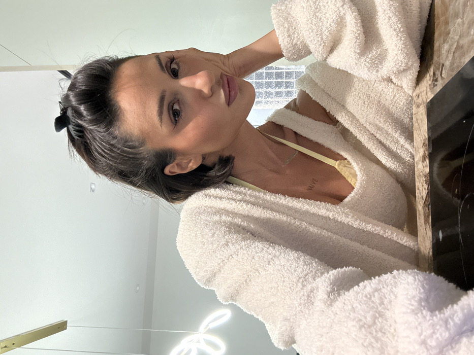 A female subject is depicted leaning forward over a dark, reflective surface, likely a kitchen countertop, in a modern indoor setting within the city of Unknown, Unknown. She appears to be in her late 20s to early 30s, with dark hair pulled back and secured by a dark claw clip. Her dark eyes are directed towards the camera, and her expression is neutral with slightly parted, glossy lips. Her left hand rests against her jawline, supporting her head. She wears a cream-colored, fuzzy, long-sleeved top or robe, which is pulled down to expose her left shoulder and collarbone. A yellow bra strap is visible on her left shoulder, and a tattoo reading "NAIVE" is present on her upper chest. A thin necklace is also visible around her neck. The background features a light-colored wall, possibly pale green, with a white abstract neon light fixture and a gold linear pendant light hanging from a white ceiling with recessed lights. To the right, a window or glass block partition with a square grid pattern is visible.