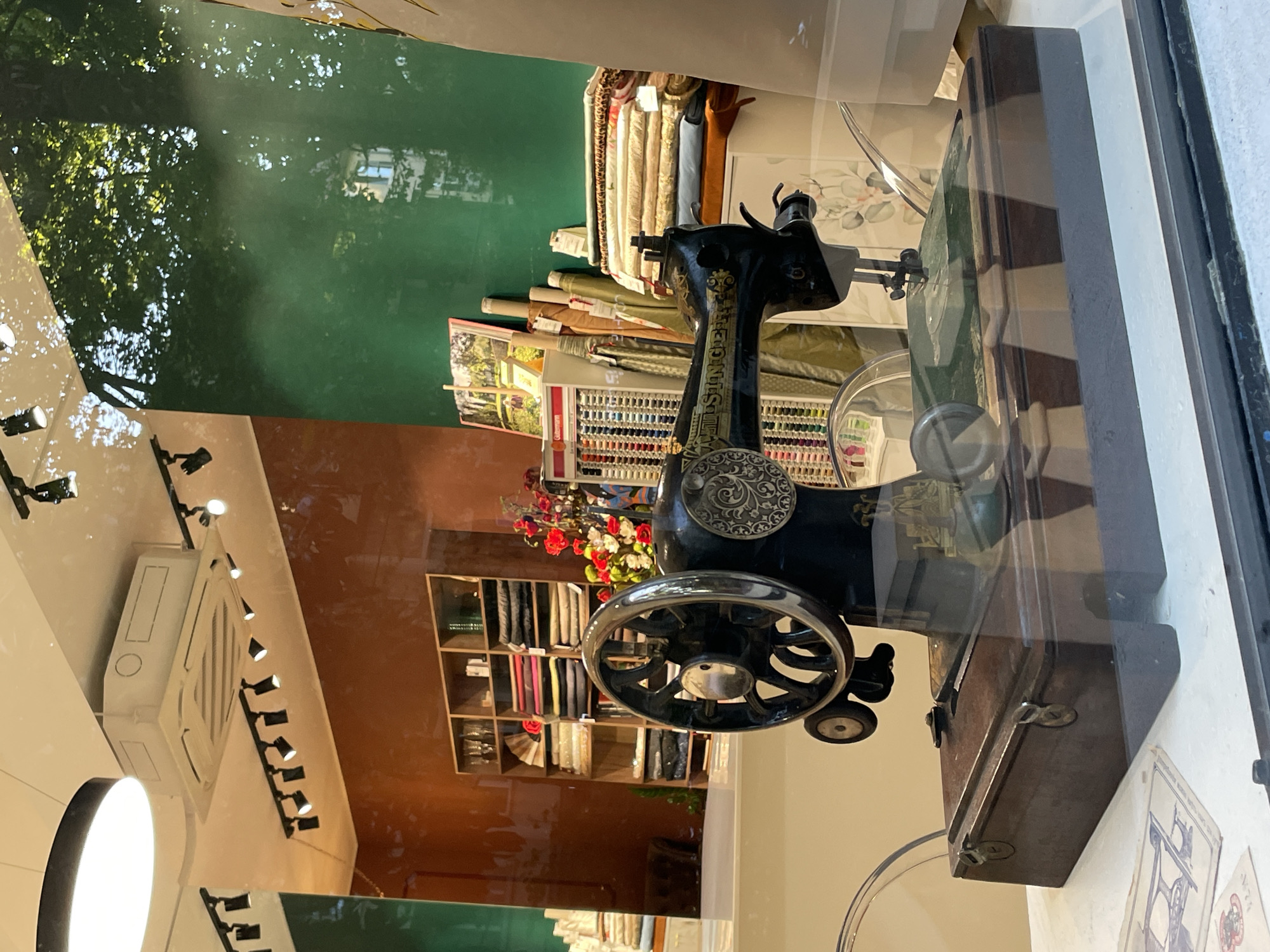 This is a vertically oriented, eye-level shot taken through a window, likely from a street, into what appears to be a fabric or sewing shop. The primary subject is an antique black sewing machine, prominently displayed. It has intricate floral-patterned metalwork and a large spoked handwheel. Next to the sewing machine, there is a wooden case. In the background, to the left of the sewing machine, is a wall painted a deep green, reflecting dappled sunlight and tree branches, suggesting it's daytime and outdoors. Further into the shop, shelves are filled with rolls of colorful fabrics and spools of thread, indicating the nature of the business. A small vase with red and white flowers sits on a shelf above a cubby-style shelving unit filled with folded fabric swatches. The overall scene is well-lit, with the sunlight creating reflections on the window. The image captures a blend of antique craftsmanship and the organized abundance of a haberdashery. The location is specified as Sochi, Russia. There is no visible text in the image.