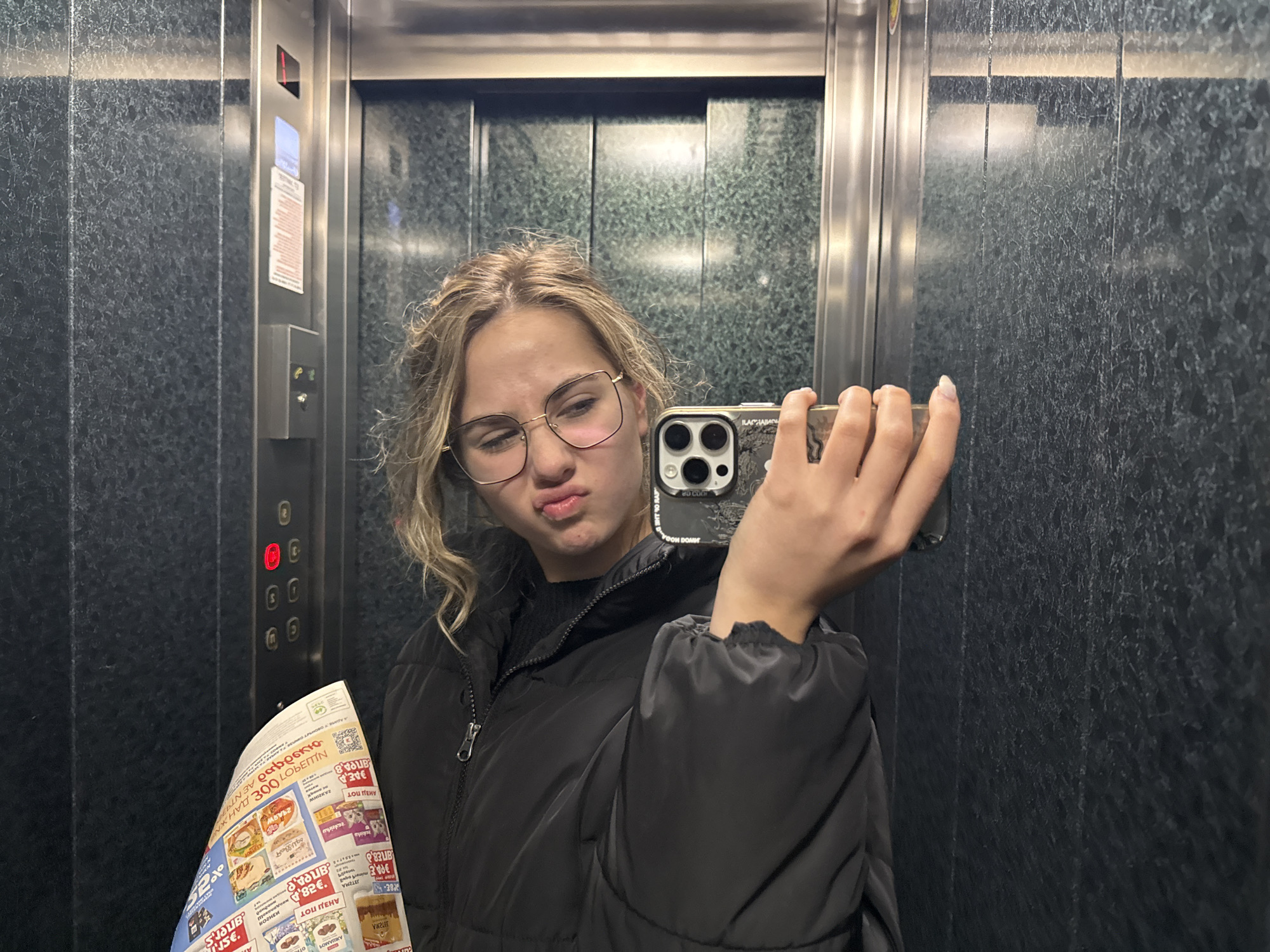A woman in an elevator in what appears to be Blagoevgrad, Bulgaria, is taking a selfie. The woman has blonde hair, glasses, and is wearing a black puffer jacket. She is holding a black smartphone in her right hand. In her left hand, she holds a Bulgarian supermarket flyer advertising food products. The elevator's control panel is visible to her left. The flyer appears to feature Cyrillic text and price tags in euros. The elevator walls are dark gray.
