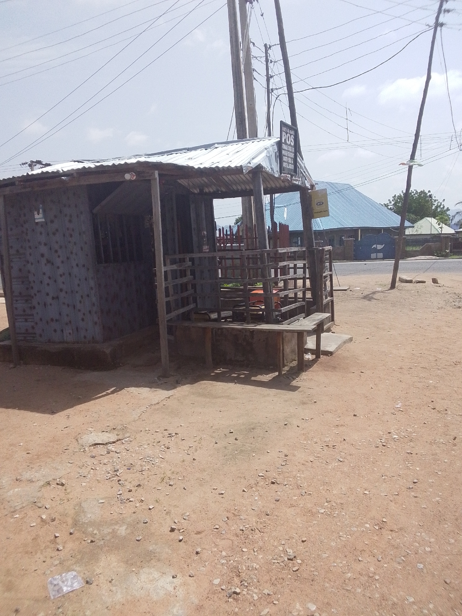 The image depicts an outdoor scene in Jalingo, Nigeria, featuring a small, weathered wooden kiosk or shop with a corrugated metal roof. The structure is situated on a dirt ground with scattered small stones and debris. A wooden bench and a railing system are attached to the front of the kiosk. A sign with "POS" is visible on the kiosk. In the background, a building with a blue, pitched roof is present, along with utility poles and overhead wires. The environment appears to be a roadside area, with indications of urban infrastructure.