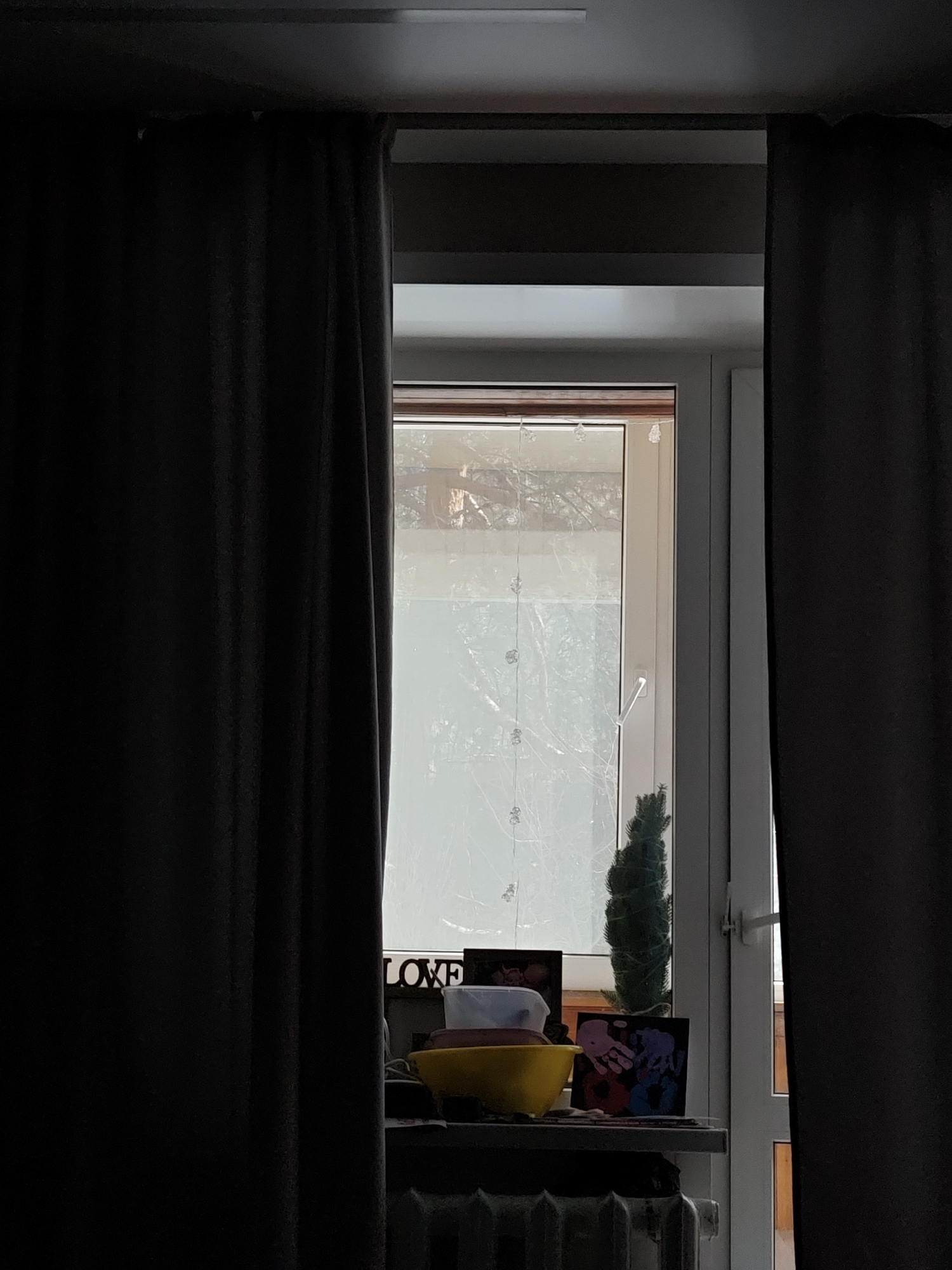 The scene depicts an interior view of a room located in Chaikovsky, Russia, observed through a window. Dark, heavy curtains, appearing to be of a blackout type, are drawn partially open to the left and right, framing a central window. The window features a modern white PVC frame with a visible white handle on the right-hand pane. A string of small, clear, possibly snowflake or star-shaped decorative lights hangs vertically across the window's interior.
The view through the window shows bright, hazy daylight illuminating bare tree branches, suggesting a colder season. On the window sill, arranged from left to right, are a dark object or sign with the letters "LOVE" partially visible, a white container, a yellow bowl, a small dark green artificial conical tree or topiary, and a framed piece of artwork displaying multiple colorful handprints (pink, purple, red, and blue). A white radiator is partially visible beneath the window sill. No individuals or active interactions are present within the depicted frame.