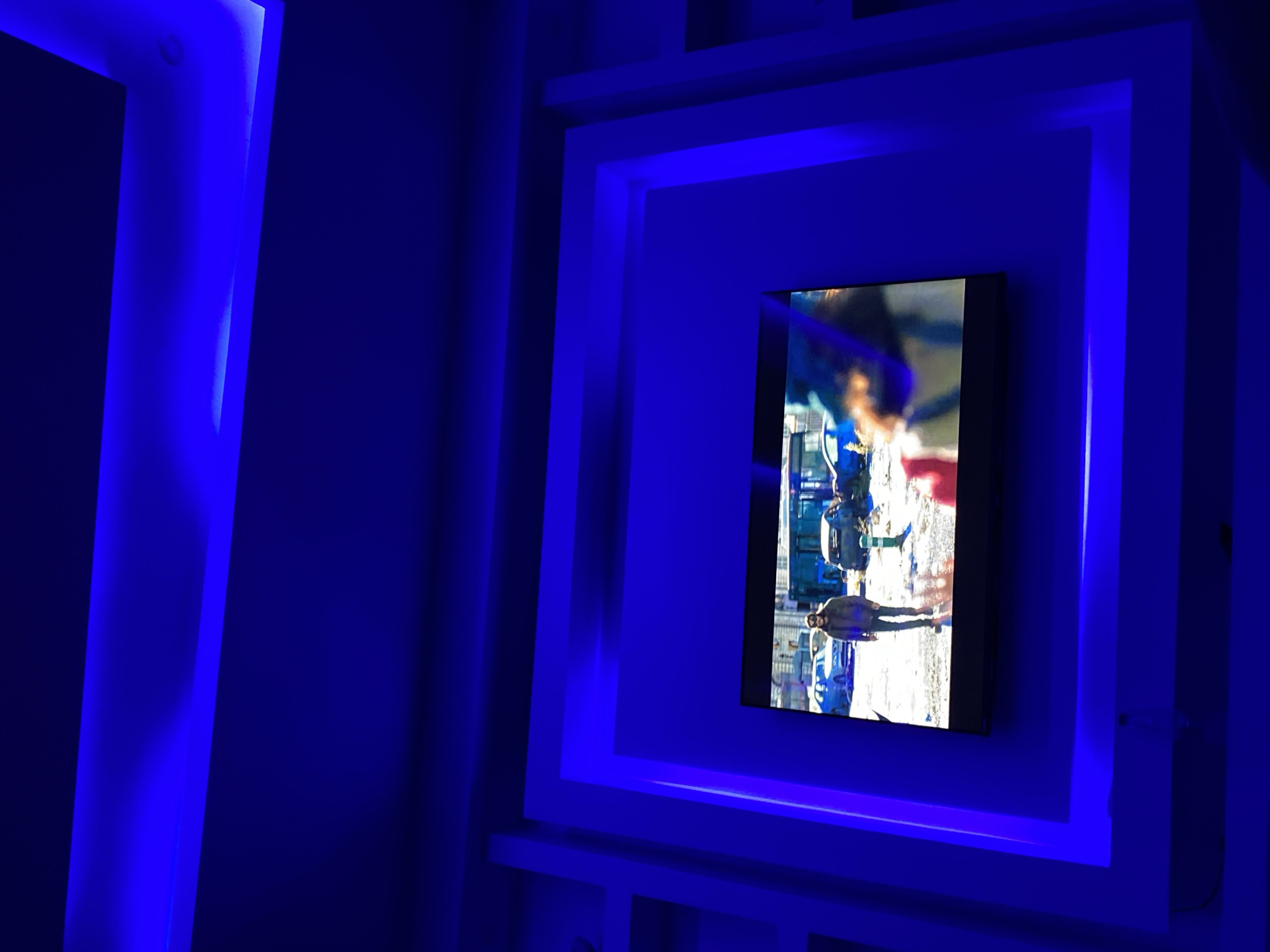 A television is mounted in a white, square frame on a wall. The television screen displays a scene with a man walking down a street in what appears to be Lagos, Nigeria, based on the presence of yellow commercial buses. The wall is illuminated with blue LED lights around the frame and in other areas. The environment is dimly lit and the blue lighting is prominent.