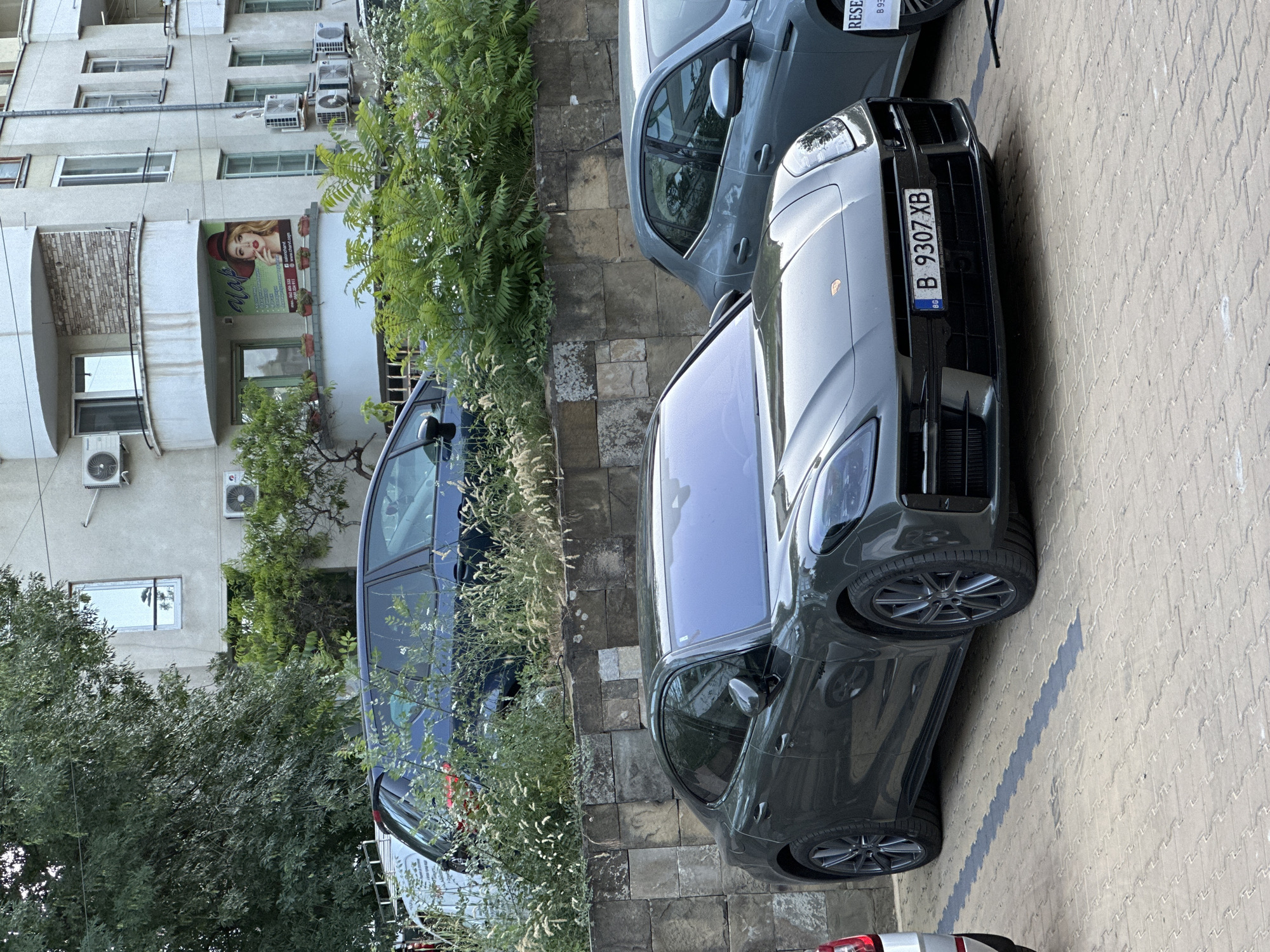 A dark gray Porsche Panamera sedan is parked in a paved lot in front of a stone wall. The license plate reads "B 9307 XB". Behind the Porsche, a dark blue hatchback is partially visible, overgrown with greenery. Further back, an apartment building with several windows and air conditioning units is visible. A large tree with dense foliage is to the left of the building. In the foreground, the textured pavement is visible, with a dark gray painted line marking a parking space. The scene appears to be in a city, likely Veliko Tarnovo, Bulgaria, given the license plate format. There are no people or animals visible in the image.