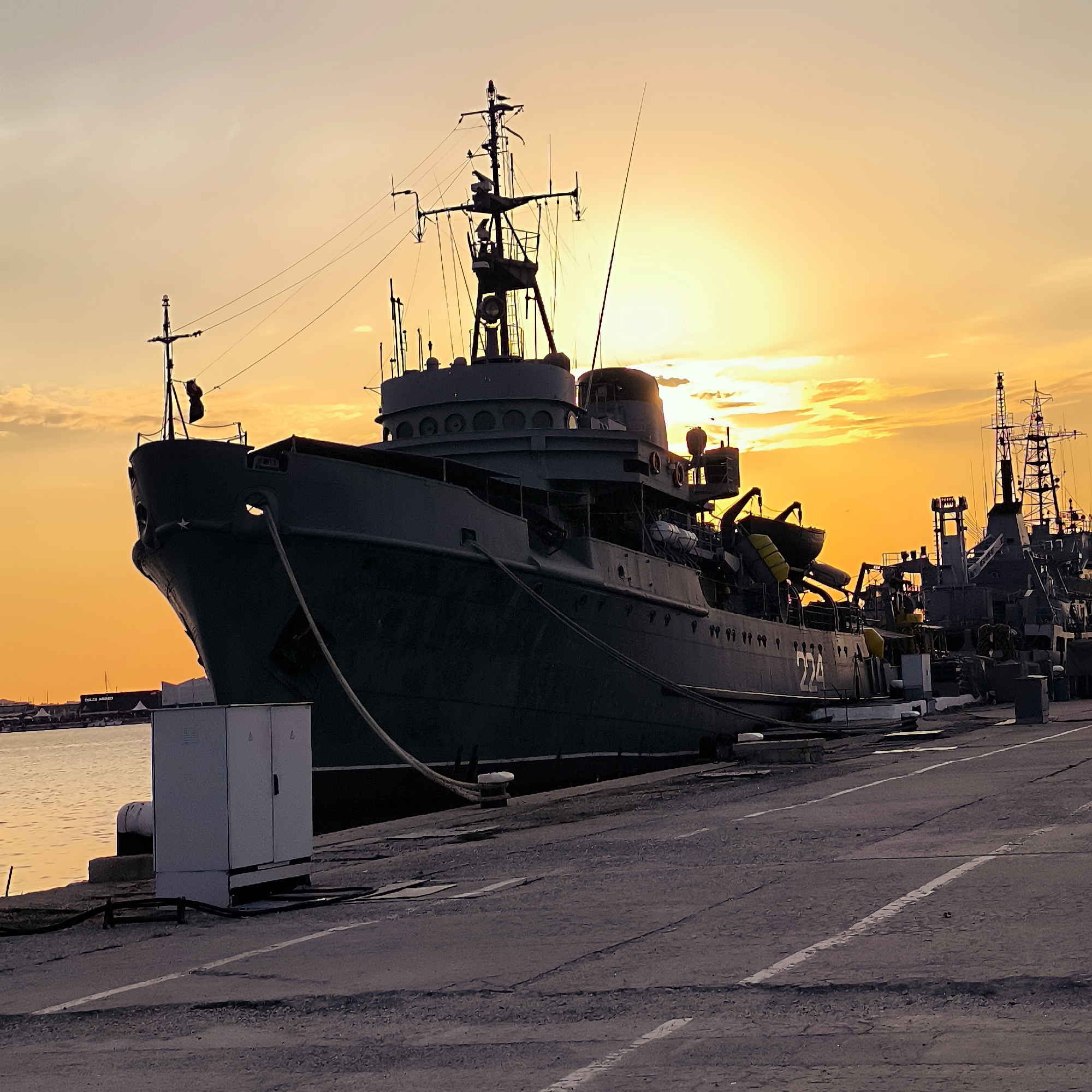 The image depicts a large, dark-colored ship docked at a pier at sunset. The sky is a vibrant orange and yellow, with some clouds present. The ship is positioned in the foreground, taking up a significant portion of the frame. It appears to be a naval vessel, with various structures and antennas visible on its deck. The pier itself is made of concrete and has some electrical boxes and bollards. In the background, other ships and structures can be seen, suggesting a port or harbor setting. The water is calm and reflects the colors of the sunset. The overall impression is one of a tranquil, end-of-day scene in a maritime environment. This scene is located in the port of Varna, Bulgaria.
