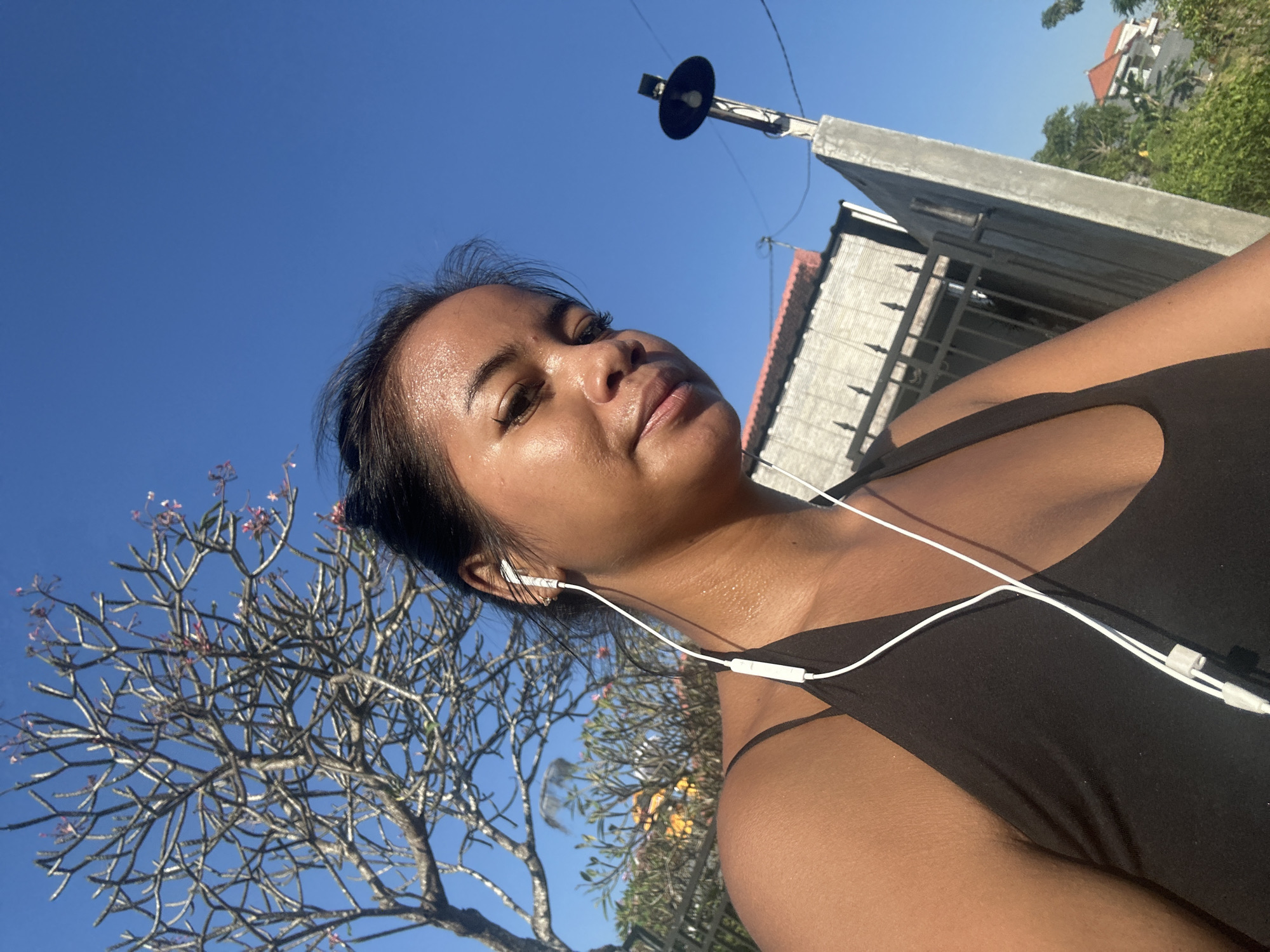 A young woman with dark hair pulled back and wearing a black tank top is positioned in the foreground of the image, with her head tilted slightly downwards. She has on white earphones, and her skin appears to be glistening with sweat. The background consists of a clear blue sky, a leafless tree with small pink blossoms, a fence, and a portion of a building. To the right, a concrete post with a light fixture is visible. The scene suggests an outdoor setting, possibly in Sukawati, Indonesia, given the architectural style and vegetation.