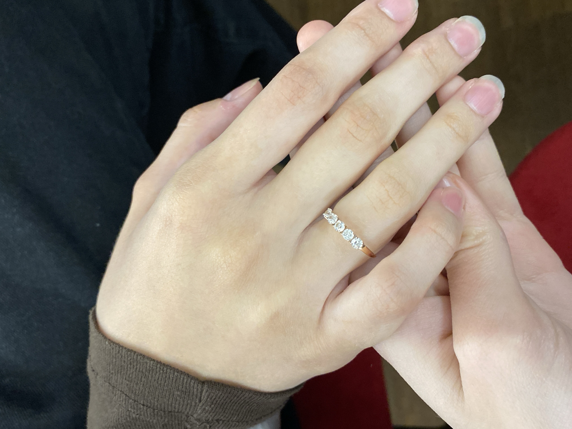 The image features two light-skinned hands clasped together, suggesting a tender or supportive interaction between two individuals, or possibly one person holding their own hands. The hand on the left, closer to the viewer, has a rose gold or gold band ring on its ring finger, set with five small, clear, possibly diamond stones. The nails on both hands are natural, appearing healthy and well-kept with a clear or light pink hue.
The hand on the left is wearing a brown, ribbed long-sleeved garment, visible at the wrist. Behind this hand, a dark, possibly black, fabric or surface is visible, suggesting clothing or furniture. To the right of the hands, a reddish or maroon fabric, also possibly clothing or upholstery, forms part of the background. The lighting is soft and even, indicating an indoor setting. There is no discernible text within the image. The overall impression is one of intimacy, connection, and focus on the hands and the significant piece of jewelry.