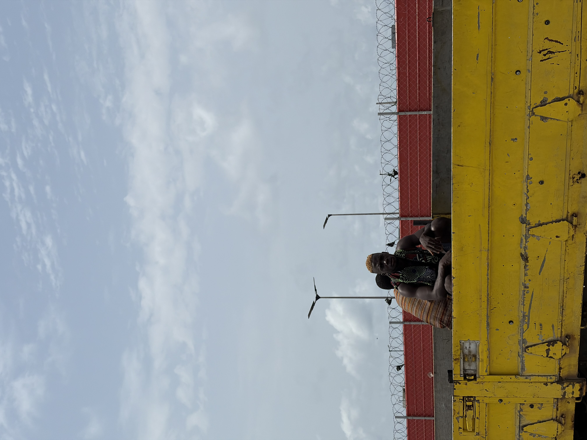 Two people are sitting atop a yellow structure in Alaguntan, Nigeria. A red wall topped with barbed wire is visible behind them, alongside lampposts. The sky is overcast with light clouds.