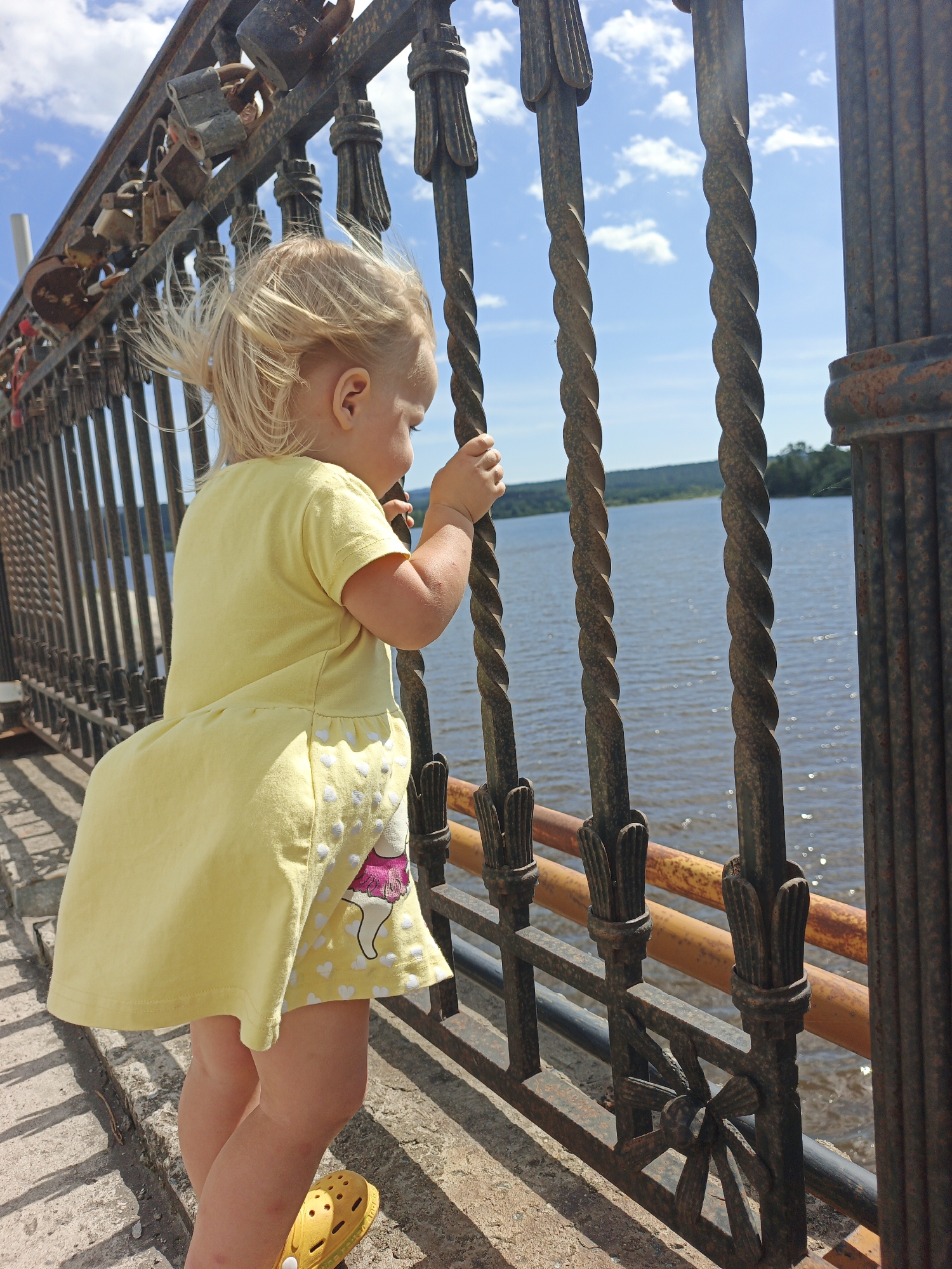 A young child, facing away from the camera, looks out at a wide body of water. The child is wearing a bright yellow dress with a subtle heart pattern and pink accents. They are standing on a concrete walkway next to a black, ornate metal railing. Attached to the railing are numerous padlocks, some of which appear rusted. Behind the child, the water stretches to a tree-lined horizon under a bright blue sky with scattered white clouds. The scene is likely the embankment or a bridge in the city of Zlatoust, Russia, given the presence of the water and the architectural style of the railing, which is typical of many public spaces in Russian cities. The child's hands grip the railing as they gaze into the distance.