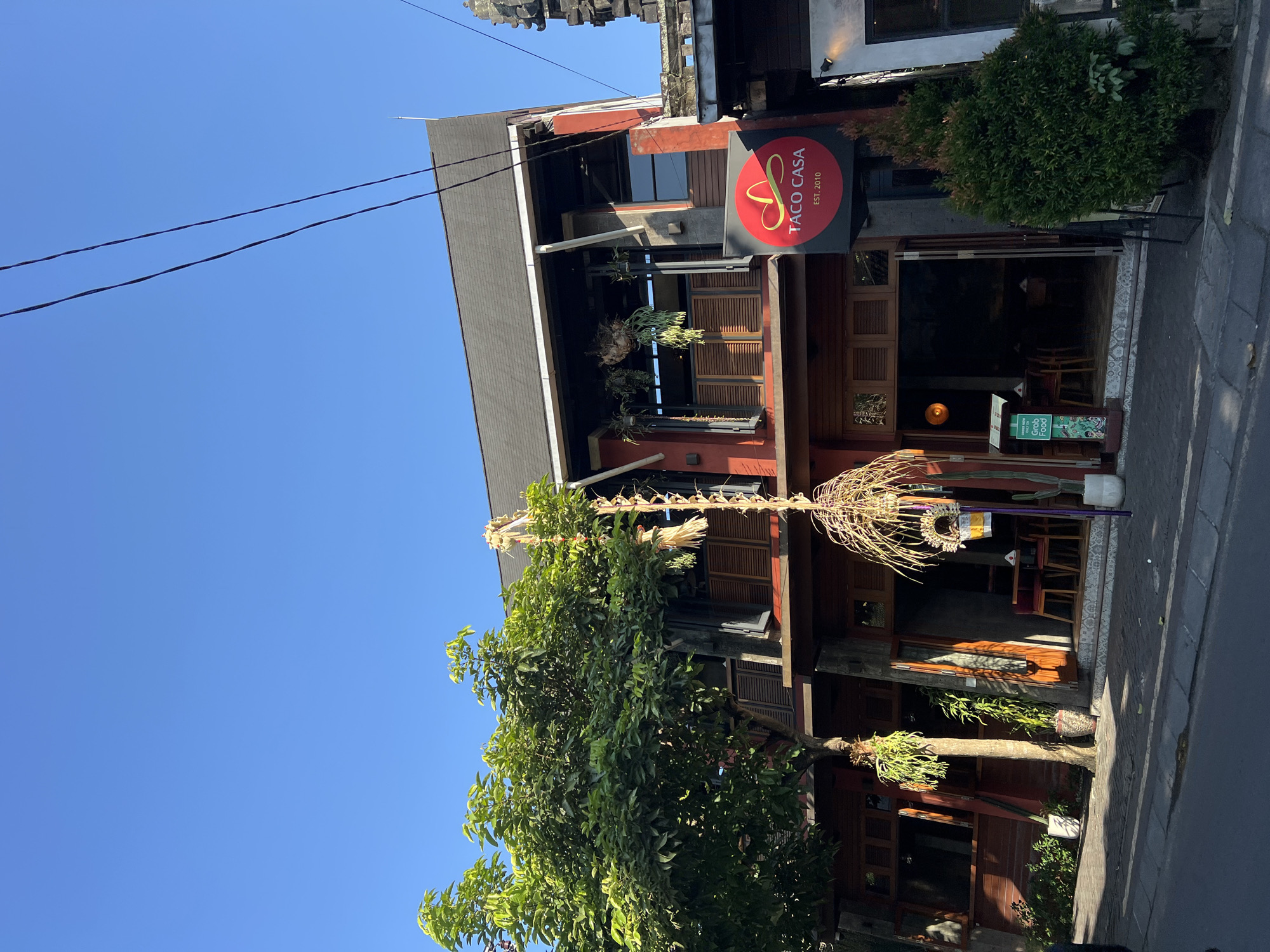 This image shows a street scene in Ubud District, Indonesia. A two-story building with a red and dark brown facade dominates the center of the frame, housing a restaurant called "Taco Casa." The restaurant has large wooden doors and windows, with tables and chairs visible inside. A red circular sign with the restaurant's name and "EST. 2010" is prominently displayed. A large green tree with lush foliage is to the left of the building, and a smaller evergreen shrub is to the right. A straw-like decoration hangs from the upper story of the building. The sky is clear and blue. The street in front of the building is paved, and a curb separates it from the sidewalk. The overall impression is that of a vibrant and welcoming establishment in a tropical setting.
