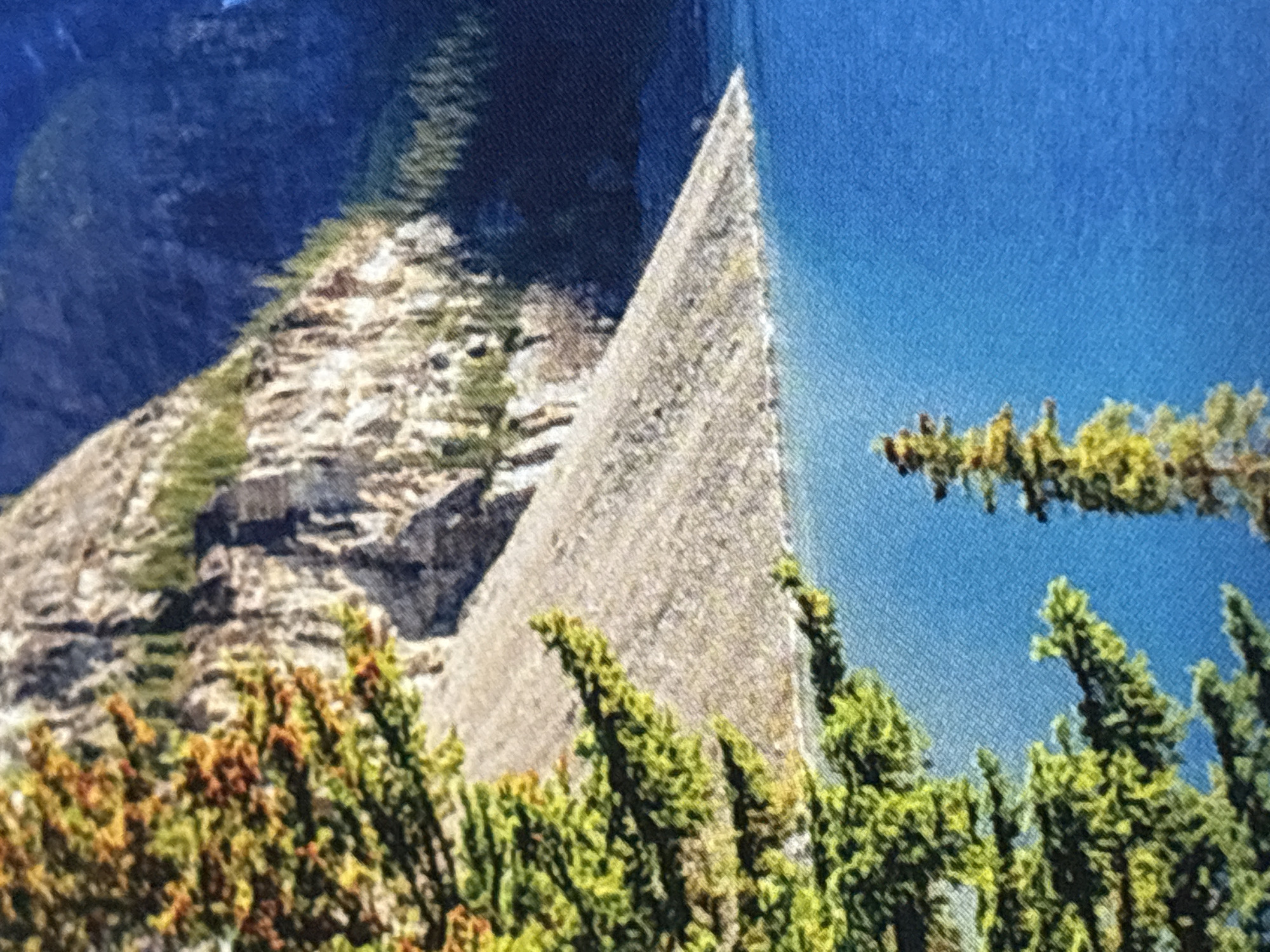 The image depicts a natural landscape with a large, triangular rock formation jutting out of a body of water. The rock has a rough, textured surface and appears to be made of light-colored stone. The water is a vibrant blue, suggesting a clear day and possibly deep water. On the left side of the frame, a steep, rocky mountainside is visible, covered in patches of green vegetation, likely trees and shrubs. These trees also appear in the foreground, with dense green foliage and some hints of brown or orange, perhaps indicating autumn colors or dry brush.
The overall scene is a natural environment, likely a mountain lake or fjord. The lighting suggests it could be daytime, with sunlight illuminating the rock and water. There are no people or man-made objects clearly visible, except for what might be a printed texture on the fabric of the image itself, which is consistent with a photograph or a printed canvas. The setting evokes a sense of wilderness and natural beauty. There is no visible text in the image.