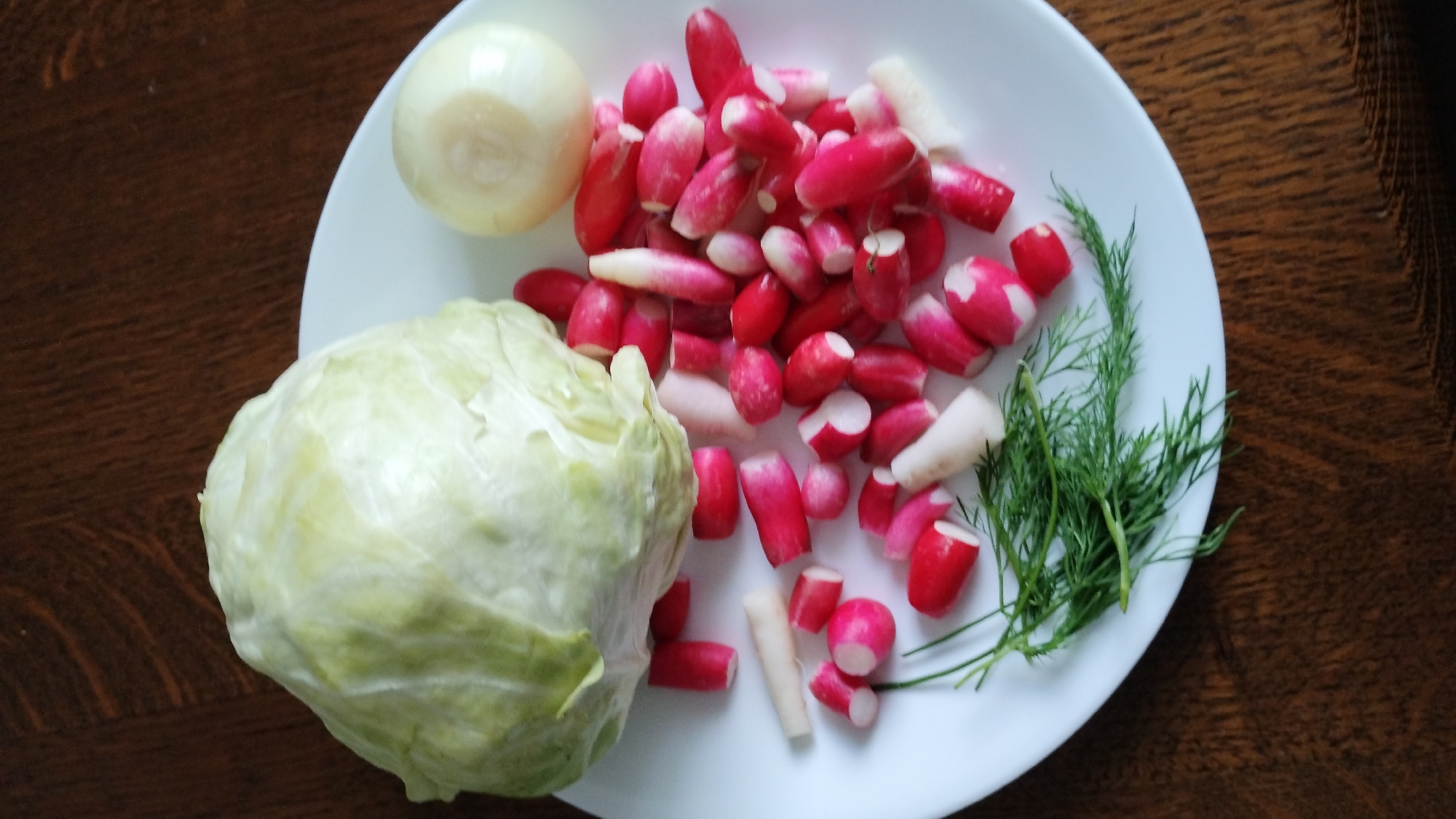 A close-up, overhead shot shows a white plate filled with chopped radishes, a half of an onion, a head of cabbage, and some dill. The plate is set on a dark brown wooden table. The radishes are sliced into small pieces, showing their pink and white interiors. The cabbage is a pale green and appears to be a whole head. The onion is a white sphere. The dill is a bright green herb, with delicate fronds. The lighting is natural and casts soft shadows. The scene is staged, likely for a meal preparation or recipe demonstration. There are no people or animals in the image.