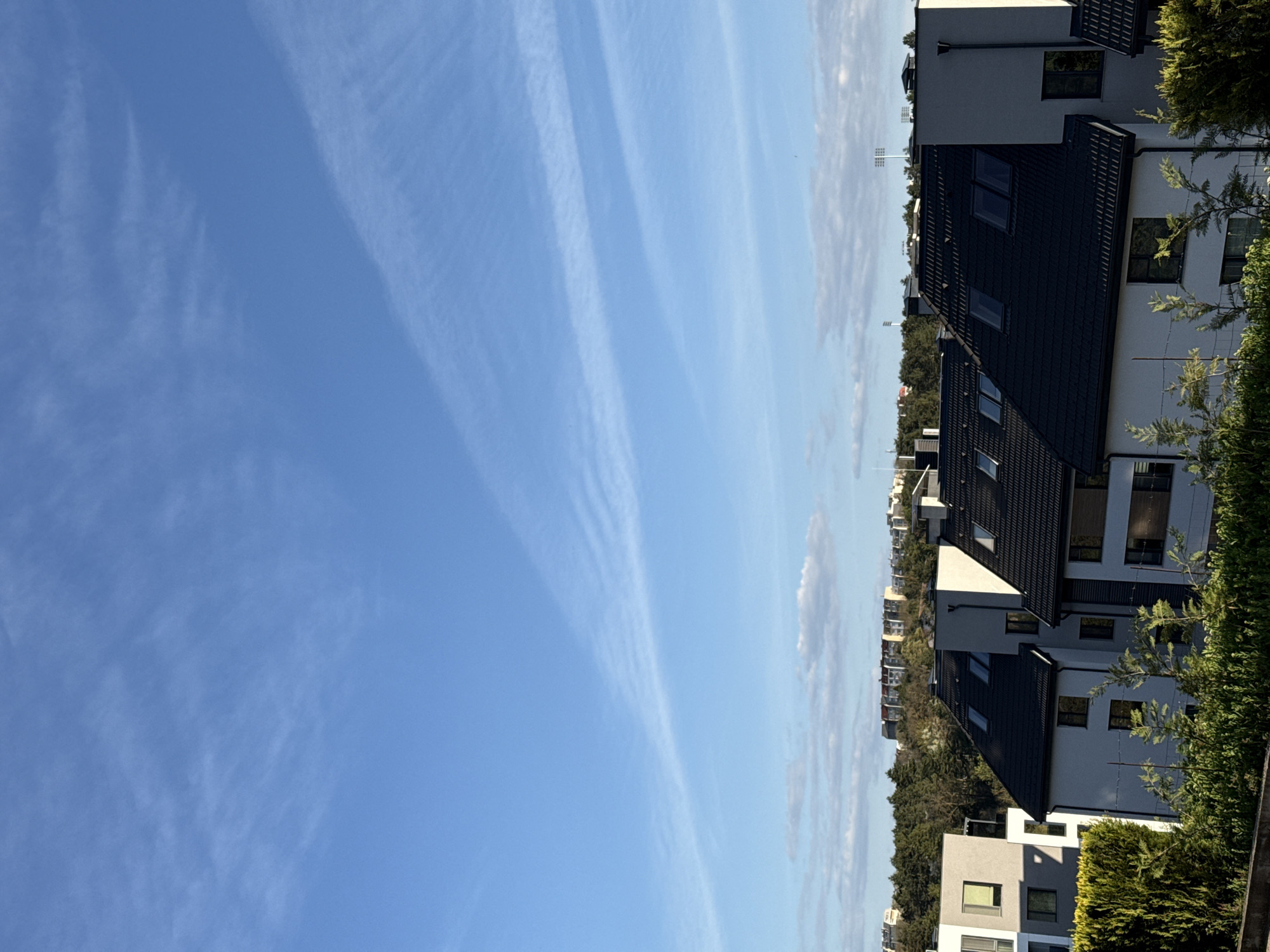 The image displays an outdoor scene dominated by a vast blue sky with some distinctive wispy clouds, likely taken in Dobrich, Bulgaria. The sky, occupying the left and upper portion of the frame (when correctly oriented), is a vibrant clear blue with long, thin, white clouds stretched across it, some appearing in parallel streaks.
To the right and bottom of the frame are several modern residential buildings, likely townhouses or detached houses. These structures feature dark, possibly black or very dark grey, pitched roofs with visible roof tiles or panels, and light-colored (white or light grey) facades. Numerous rectangular windows, many with dark frames, are visible on the buildings' walls and roofs. Green foliage, including tall trees and dense bushes or hedges, is prominent in the immediate foreground on the right and also along the line of houses, providing natural landscaping.
In the far distance, on the horizon line between the buildings and the sky, several faint, tall structures, possibly stadium lights or communication towers, can be discerned. The overall lighting suggests bright daylight, indicating fair weather with clear visibility. No specific activities are taking place, and no people or visible text are present in the image.
