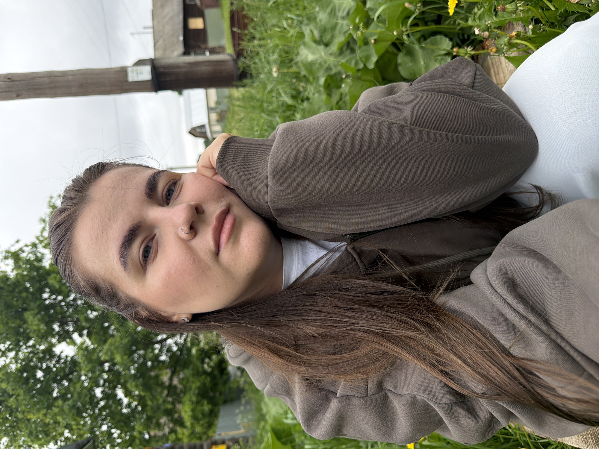 A portrait-style photograph, rotated 90 degrees clockwise, captures a young person with long, dark brown hair looking directly at the camera with a calm and relaxed expression. They are wearing a dark brown or khaki-colored hooded sweatshirt over a white t-shirt, with their left hand gently supporting their cheek. Notable details on the subject include a nose piercing on the left nostril and a small earring in their left ear.

The scene is set outdoors in Marakushi, Russia, under an overcast sky, indicating a cloudy daylight. The background features abundant green foliage, including various plants and what appears to be grass, with small yellow flowers visible on the right side. A prominent wooden utility pole, possibly with a small, illegible sign attached, stands vertically in the left background. Further back, blurred wooden structures and hints of buildings suggest a rural or semi-rural environment. No specific activity beyond posing for the photograph is depicted. The overall mood is tranquil. There is no legible text visible in the image.