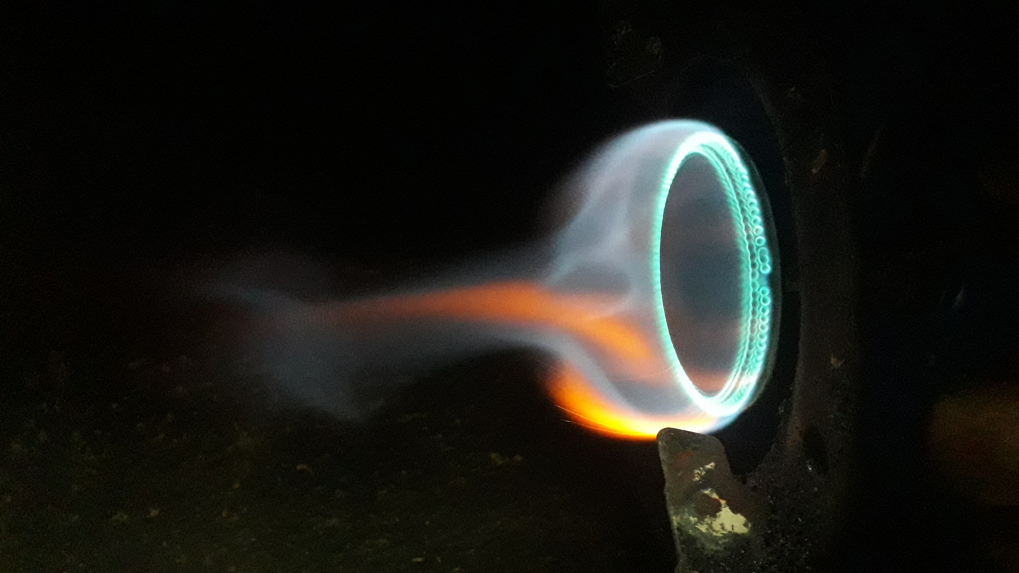 A close-up image shows a lit gas burner. The burner head is circular with a ring of blue flames. Emanating from the flame, there's a plume of orange-yellow heat, diffusing into a wisp of smoke. The burner is dark and metallic. The background is dark and out of focus. There is no indication of this image being taken in Nkpor, Nigeria.