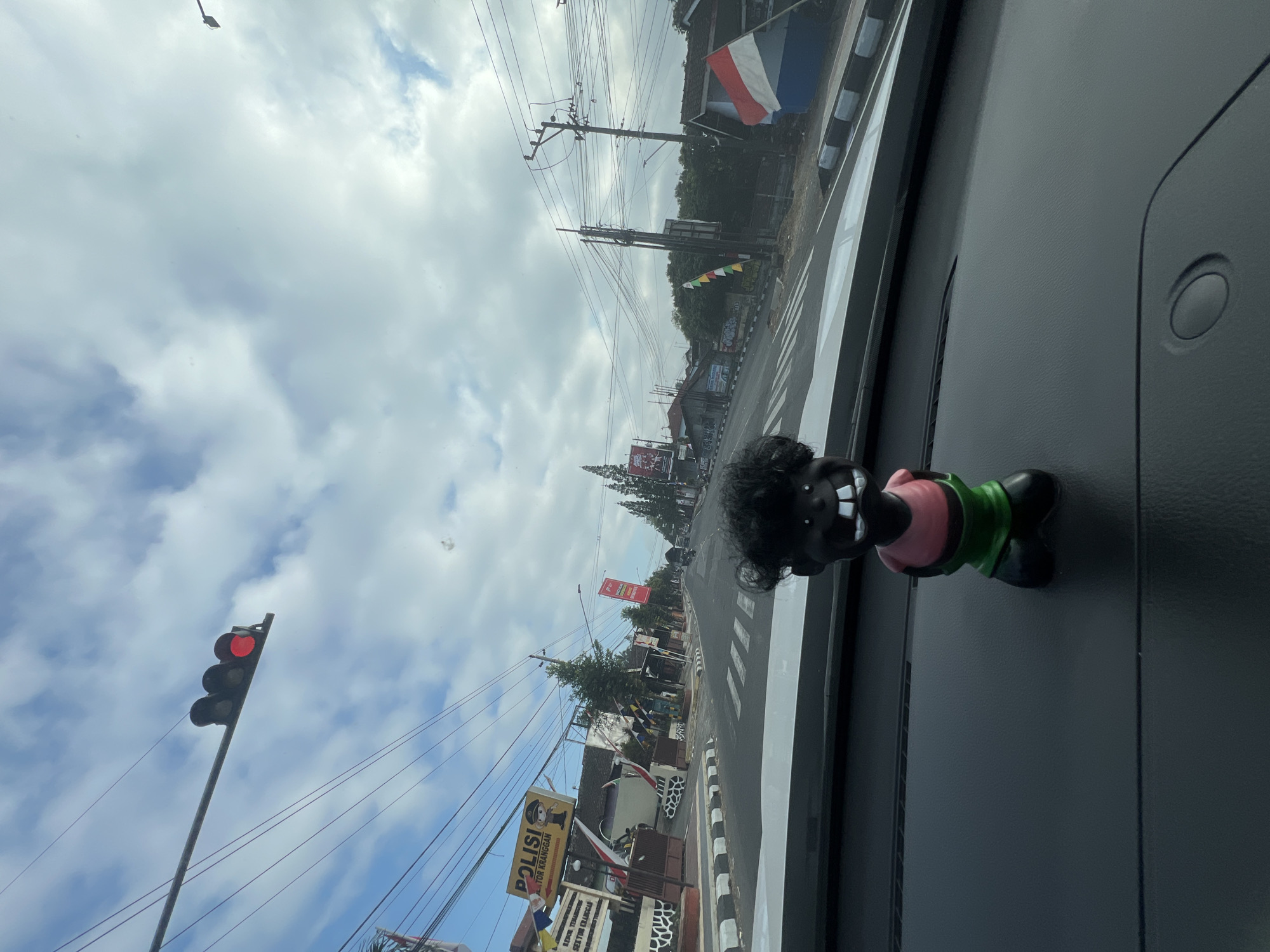 An interior view from a vehicle shows a doll with black curly hair, a pink shirt, and green shorts on the dashboard. Outside, a street scene in Kranggan, Indonesia, unfolds under a cloudy sky. A traffic light with a red signal is visible overhead. Buildings line the street, including one with a sign reading "POLISI SEKTOR KRANGGAN." Indonesian flags, one tricolor, are displayed. Power lines crisscross the sky. The street has pedestrian crosswalks and appears to be a busy urban area.