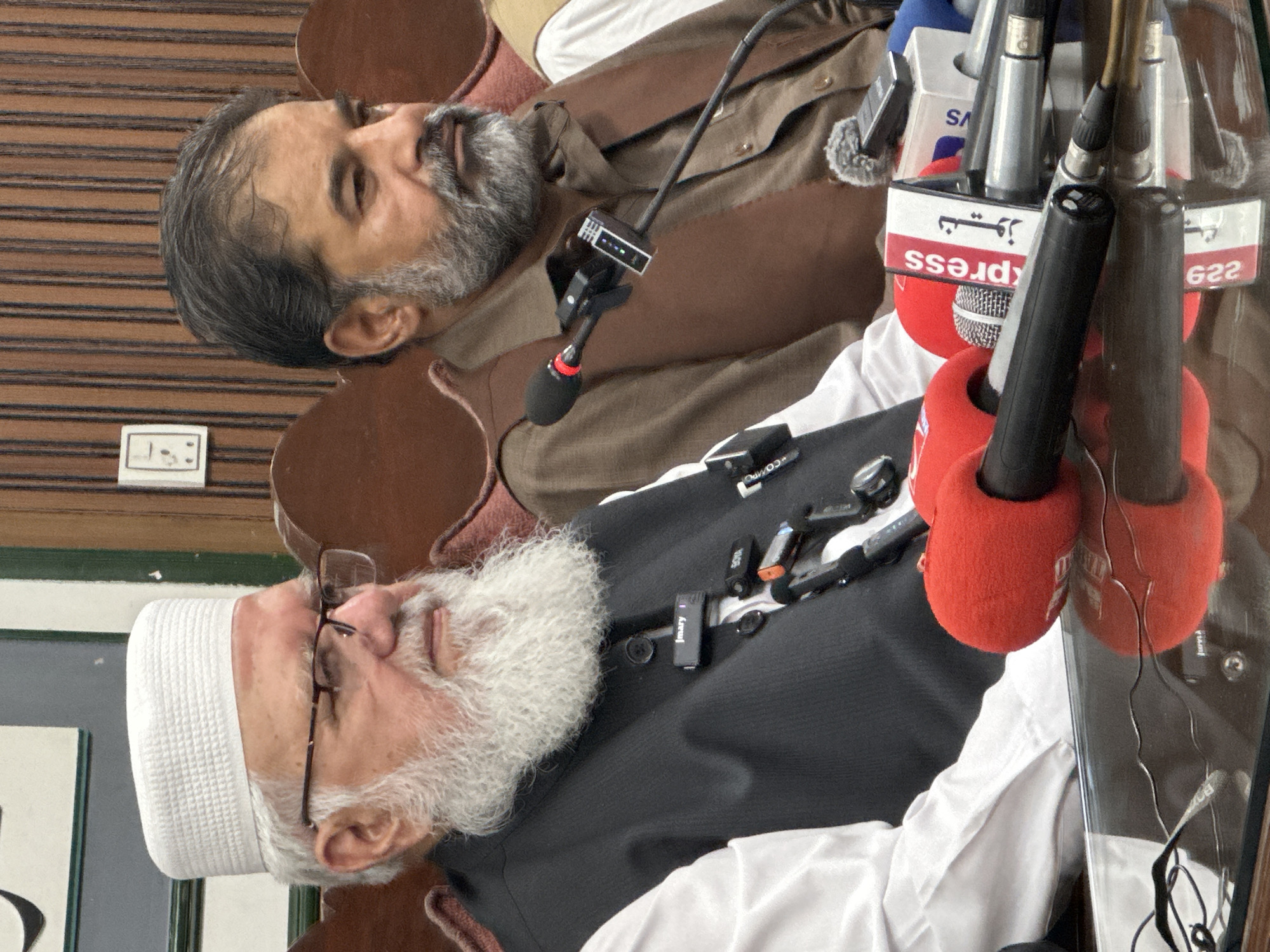 Two men are seated at a table in what appears to be a press conference. The man on the left is older, with a white beard and cap, wearing a black vest over a white shirt. He has a microphone clipped to his vest. The man on the right has a grey beard and is wearing a brown tunic and vest. Both men are looking to their right. In front of them on the table are several microphones, some with red foam covers, and a white box with the word "Express" and Urdu script on it. The background consists of wooden paneling and upholstered chairs. The setting is likely a government building or conference room in Quetta City Tehsil, Pakistan, given the presence of microphones and the "Express" branding which could indicate a local media outlet.