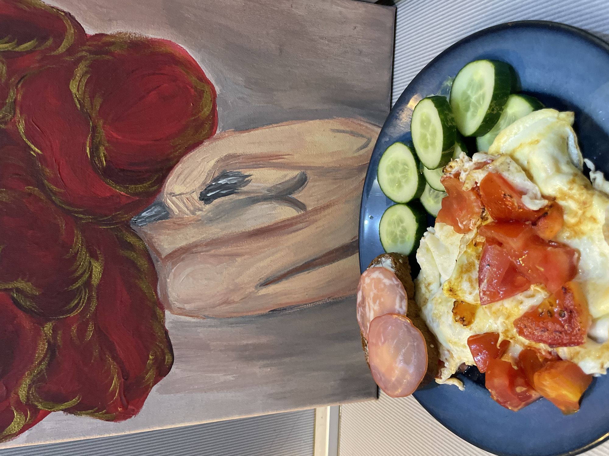 This is a photograph of a plate of food in front of a painting. The plate, which is dark blue, contains a fried egg with chopped tomatoes and several slices of cucumber. There is also a piece of bread topped with butter and two slices of ham or salami. The painting behind the plate depicts a woman's upper body and head with large, red, curly hair. The background of the painting is a pale, muted color. The setting does not provide any specific visual cues to determine its location in Sochi, Russia.