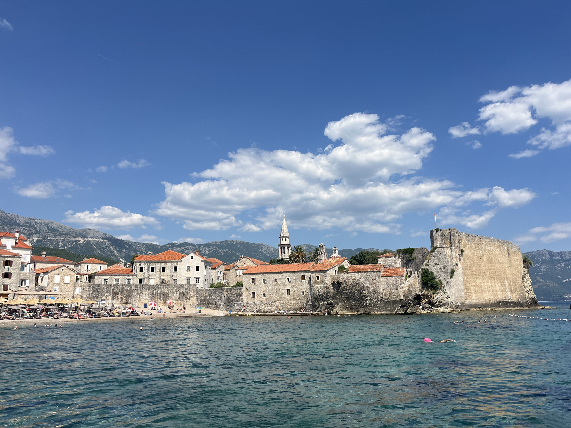 This image captures the historic Old Town of Budva, Montenegro, on a bright, sunny day. The foreground is dominated by the turquoise waters of the Adriatic Sea, with ripples and gentle waves indicating a calm surface. Several people are visible swimming in the water, including one person with a pink flotation device.
The middle ground features a bustling beach area with numerous beach chairs and umbrellas set up. People are scattered across the sand, some sunbathing, others walking or sitting. Behind the beach, the stone walls and red-tiled roofs of Budva's Old Town rise up. The architecture is characteristic of coastal Mediterranean towns, with weathered stone buildings and narrow windows. A prominent bell tower with a pointed spire rises above the rooftops, a distinctive landmark of the town.
Further back, a large, imposing stone fortress stands on a rocky outcrop overlooking the sea, its battlements and thick walls suggesting a long military history. The fortress has a commanding presence, with a flag flying from its highest point.
The background consists of a range of rugged, green-covered mountains that frame the cityscape. The sky above is a vibrant blue, dotted with fluffy white clouds, typical of a clear summer day. The overall scene is one of a popular tourist destination, combining historical architecture, a lively beach, and a beautiful natural setting.