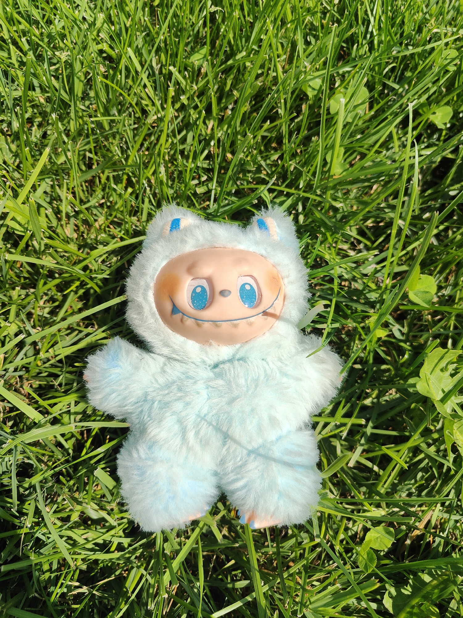 A light blue, furry plush toy is lying on a bed of green grass. The toy has a beige, round face with large blue eyes that have black pupils. The toy has a wide, smiling mouth with visible teeth and a small pink accent on its cheek. It is wearing a light blue furry hood with two small blue triangular ears on top. The toy is positioned in the center of the frame, with its head slightly tilted to the left. The grass is well-lit by sunlight, casting shadows and highlighting the texture of the blades. There are a few small green clover leaves interspersed within the grass. The location is an outdoor grassy area, consistent with a park or lawn in the city of Chornomorsk, Ukraine.
