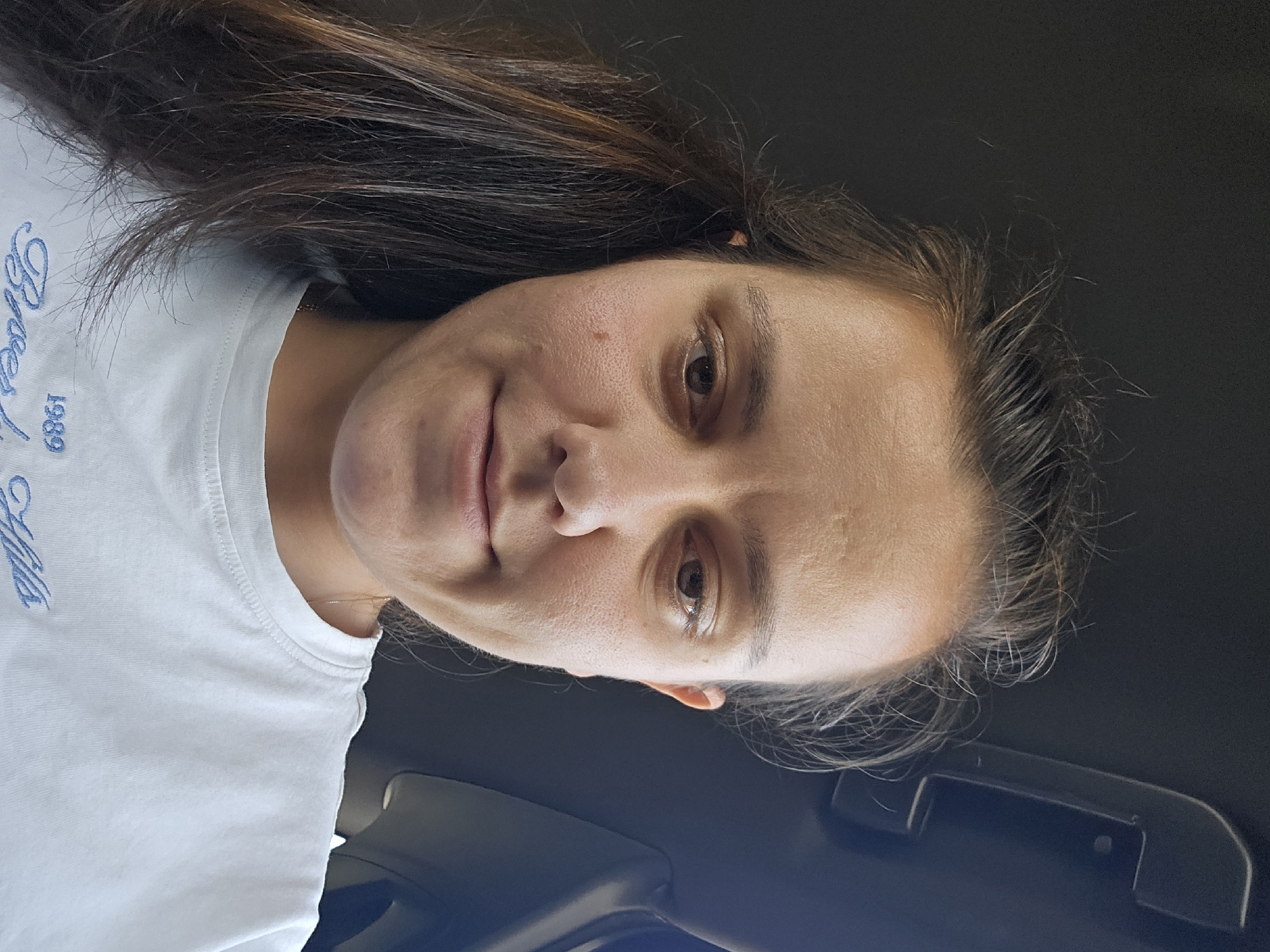 A close-up, low-angle selfie shows a woman with dark brown hair in a white t-shirt. She is looking down at the camera, and her face is partially illuminated by natural light, casting shadows on her features. Her hair is pulled back, with some strands falling loosely around her face. She is inside a vehicle, with the dark interior visible in the background. The t-shirt she is wearing has a light blue embroidered design with the text "1989" and what appears to be a name that is partially obscured. The scene appears to be casual and informal.