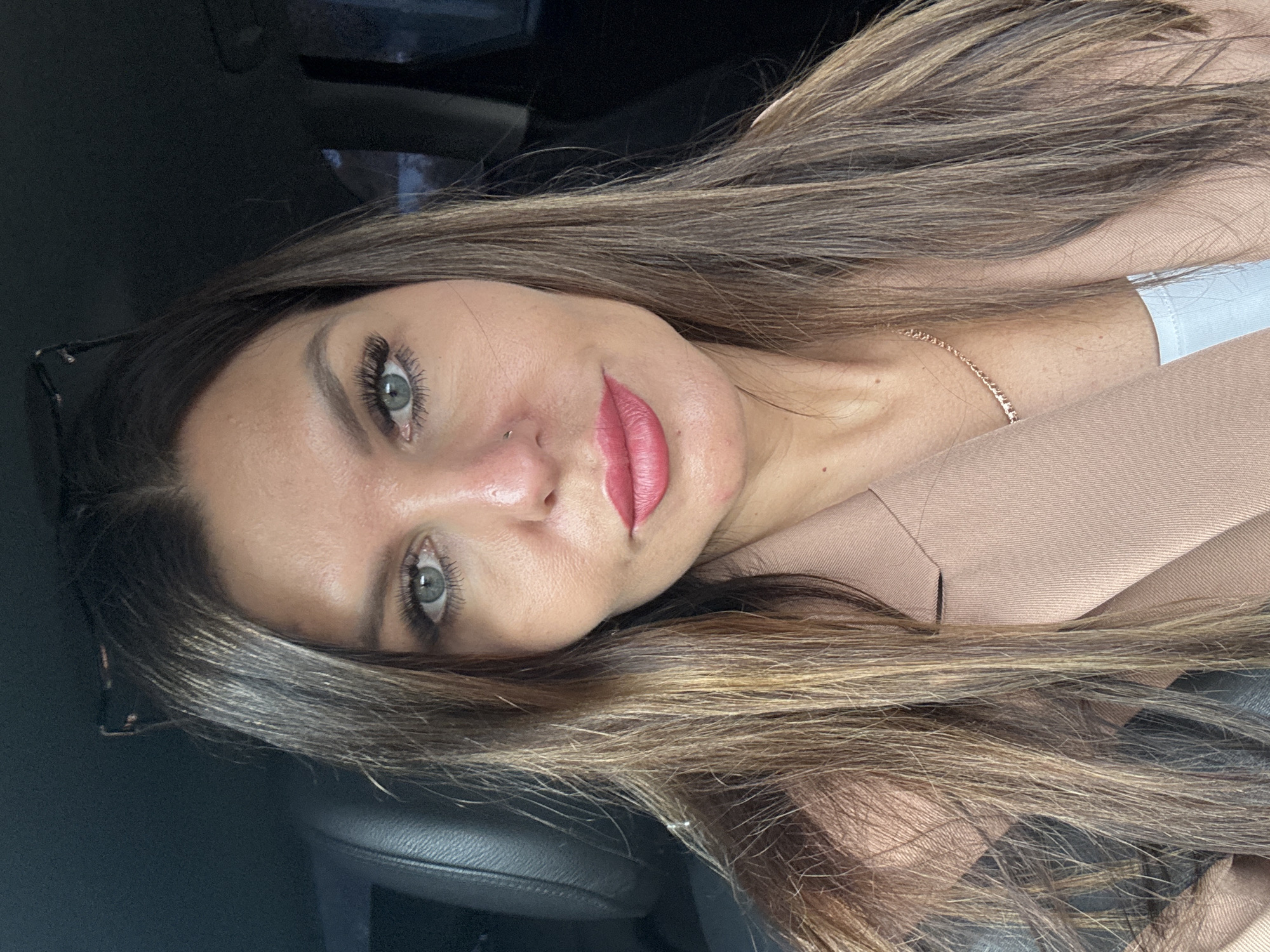 A woman with hazel eyes and long brown hair is wearing a pink blazer. She has a nose piercing and is wearing lipstick. A gold necklace is visible around her neck. She appears to be sitting in a car. The exact location within Chaikovsky, Russia, cannot be determined from the given image.