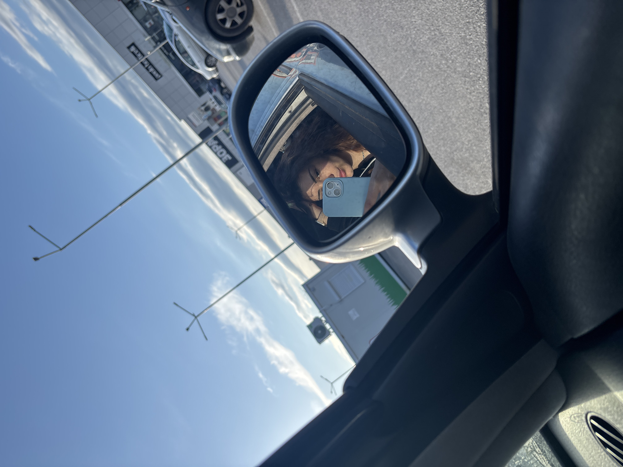 The image, taken from inside a car, shows the passenger side mirror reflecting a woman holding a phone in front of her face. The mirror also reflects a building, street lights, a car, and the sky. The interior of the car is visible in the foreground, with the dashboard and air vent partially in view. The building in the reflection has signs in Cyrillic, specifically the word "JOPA," suggesting the location may be in a region where Bulgarian is spoken, possibly Sofia, Bulgaria.