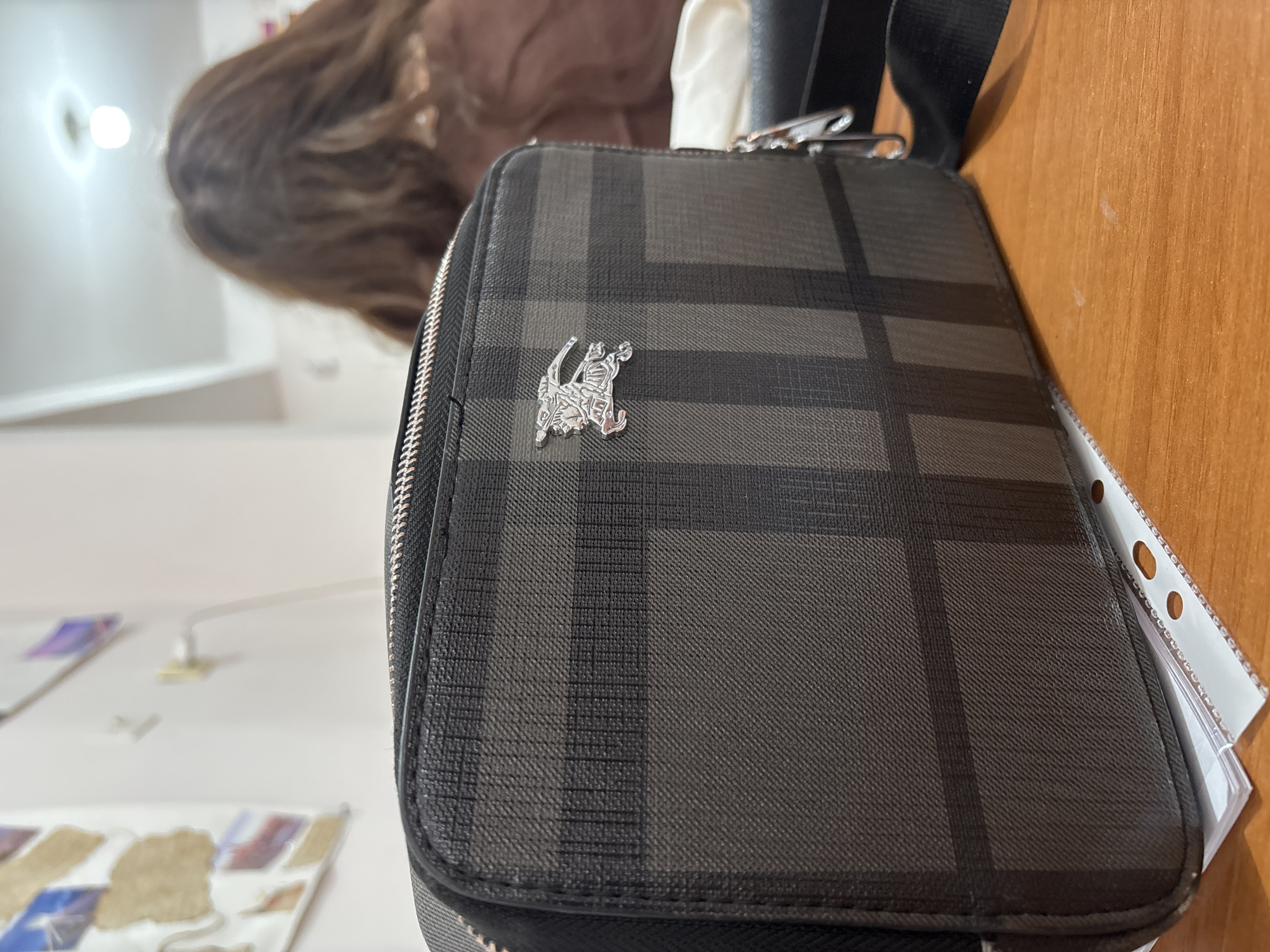 Here's a description of the image:
The image shows a Burberry crossbody bag placed on a wooden surface. The bag has a dark gray and black plaid pattern with a silver Burberry logo in the center. A portion of a person's hair is visible blurred in the upper left corner of the image. A light-colored wall is visible in the background, along with some blurry images on a table. A piece of paper with holes punched in it is partially visible near the right edge of the image. The location of Yerevan, Armenia, cannot be confirmed from this image alone.