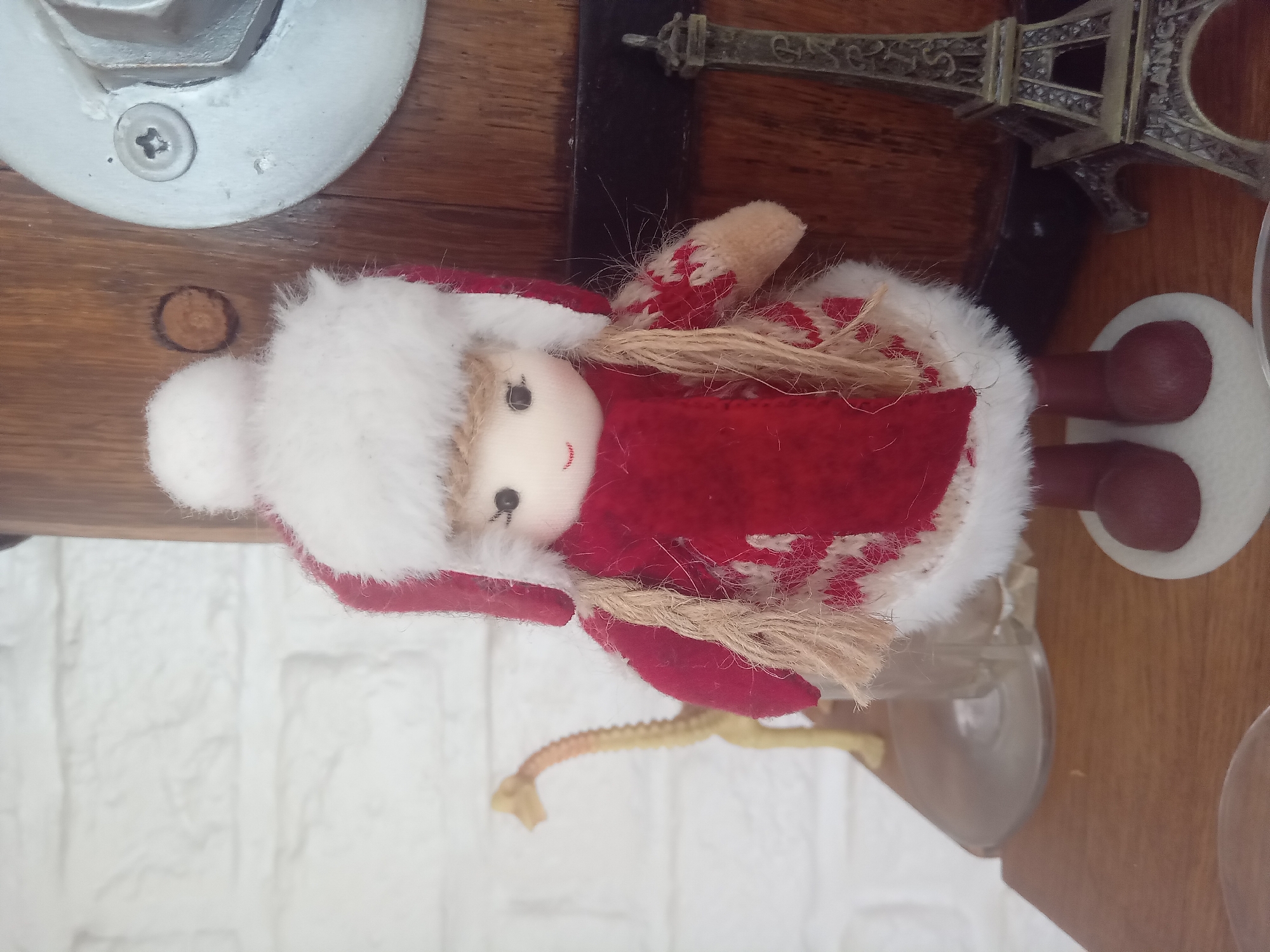 A close-up, slightly angled shot captures a small fabric doll dressed in winter attire, standing on a wooden shelf. The doll has a light-colored face with small black eyes and a stitched red smile. It wears a red hat with a white fur trim and a matching white pom-pom on top. A red scarf is draped around its neck, and it's dressed in a red coat with white snowflake patterns and white fur trim. The doll's hair is made of light brown yarn braided into two pigtails.
Behind the doll, to the left, is a blurred, light-colored brick wall. To the far left and slightly out of focus, a small, yellow toy giraffe stands on a clear glass object. To the right of the doll, a dark wooden surface is visible, possibly part of a larger object or furniture. A small, metallic Eiffel Tower replica stands on this surface. The overall lighting is soft, suggesting an indoor setting.
The image's metadata indicates it was taken on December 24, 2021, at 11:32 AM, with a Samsung SM-A525F camera. The GPS coordinates provided place the location in Terebovlia, Ternopil Oblast, Ukraine.