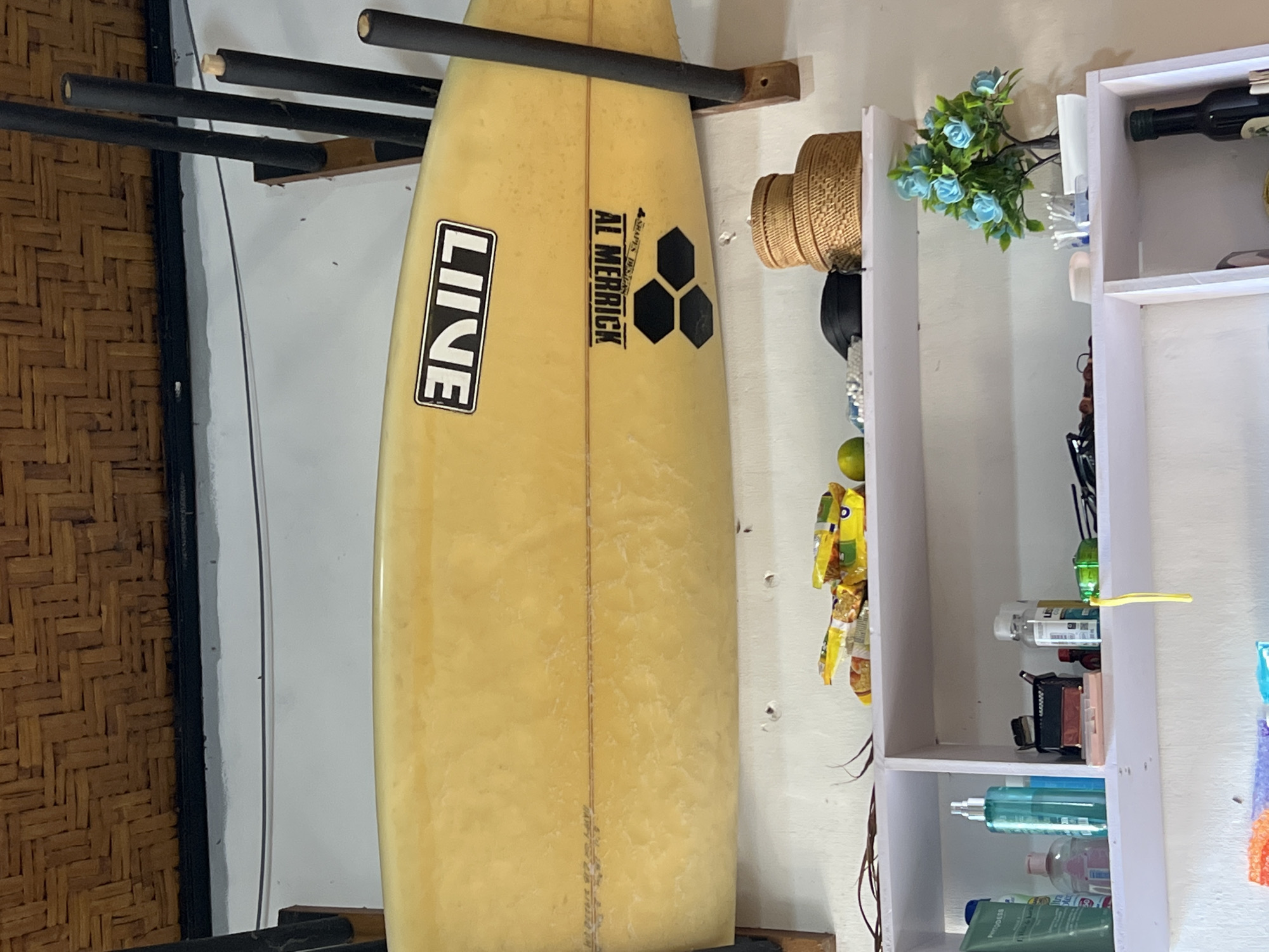 A yellow surfboard with black logos "LINE" and "AL MERRICK" is mounted on a wall rack in what appears to be a storage area or casual living space. Below the surfboard are two white shelves. The top shelf holds various items, including snacks in plastic packaging, a green lime, a black bag, and two woven baskets. The bottom shelf contains a collection of personal care items such as bottles of lotion and spray, makeup, and a small plant with blue flowers in a white pot. In the lower section of the image, a black bottle is visible on a shelf. The background consists of a woven bamboo ceiling and a white wall. The setting is likely a home in Denpasar, Indonesia, given the items and general ambiance. There are no people visible in the image.