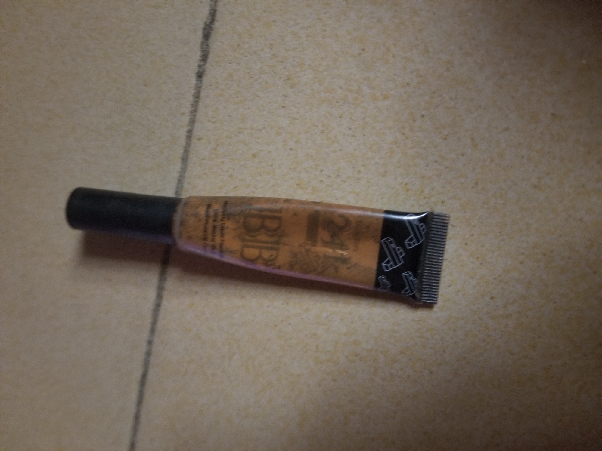 A close-up, angled shot shows a tube of BB cream on a light-colored, textured surface, possibly a tiled floor. The tube is orange with black lettering and a black cap. The text on the tube appears to read "BB" prominently, with smaller text below, including what looks like "Moisturizing Lotion" and "100% Natural." The surface has faint gray lines that suggest grout or markings. The lighting casts subtle shadows. The location is Jauro Yinu, Nigeria. There are no people or actions visible in this image.