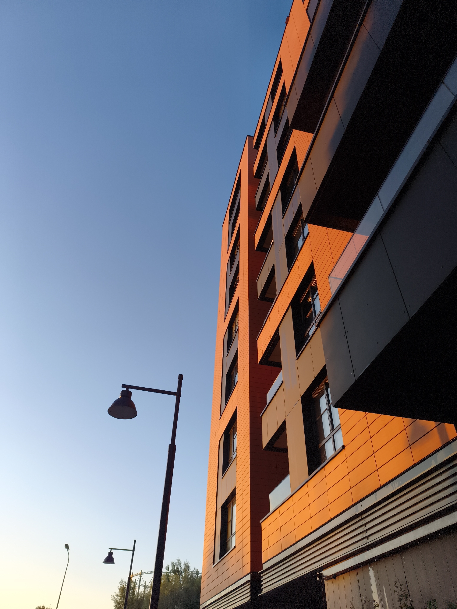 The image depicts a modern residential building in Gdańsk, Poland, bathed in the warm light of what appears to be either sunrise or sunset. The building's facade features a combination of orange and dark grey panels, with balconies jutting out. The clear blue sky occupies a significant portion of the frame, providing a stark contrast to the building's warm tones. Two streetlights are visible in the foreground, one larger and closer than the other, both silhouetted against the sky. In the distance, some trees and possibly construction equipment can be seen, indicating an urban or suburban setting. The perspective is low-angle, looking up at the building from street level.