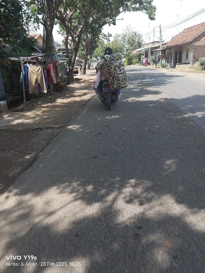 This image depicts a street scene in Patrol, Indonesia, captured on a bright, sunny day, specifically February 20, 2025, at 10:25 AM. The primary subject is a black scooter, seen from behind, traveling away from the viewer on an asphalt road. On the scooter, one person is visible, with another potentially obscured behind a very large, bulky item wrapped in patterned fabric (black with gold/cream swirls), which is loaded onto the rear of the vehicle, extending high above the rider's head.
The road surface is asphalt, appearing somewhat worn in places, and is heavily dappled with shadows cast by numerous large trees lining the street. To the left of the road, there's an unpaved dirt and gravel shoulder. Adjacent to this, a makeshift structure supports a rack displaying various items of clothing, including pants and shirts, possibly indicating a roadside vendor or laundry drying area. Behind these, simple, small buildings are nestled among dense green foliage. To the right of the road, further in the distance, more residential-style buildings with reddish-brown tiled roofs and power lines are visible under the clear sky.
The scene conveys a sense of daily life and transportation in a rural or semi-urban Indonesian setting. The scooter's license plate "E 2785 G" is visible. A watermark at the bottom left corner reads "vivo Y19s Akhtar & Arvan 20 Feb 2025, 10.25".
